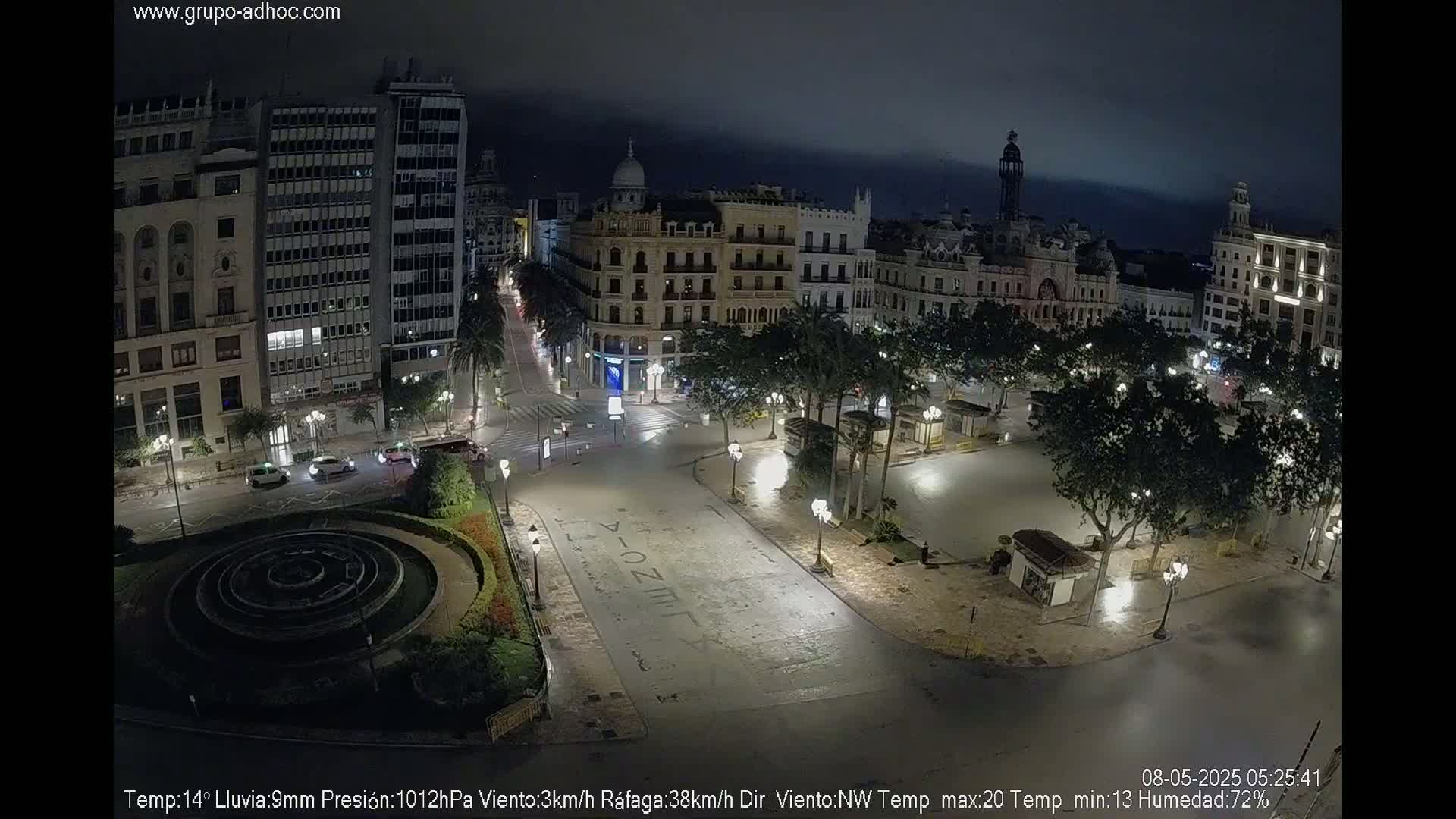 A nighttime view of a city square with a fountain, surrounded by buildings and palm trees under rainy conditions.