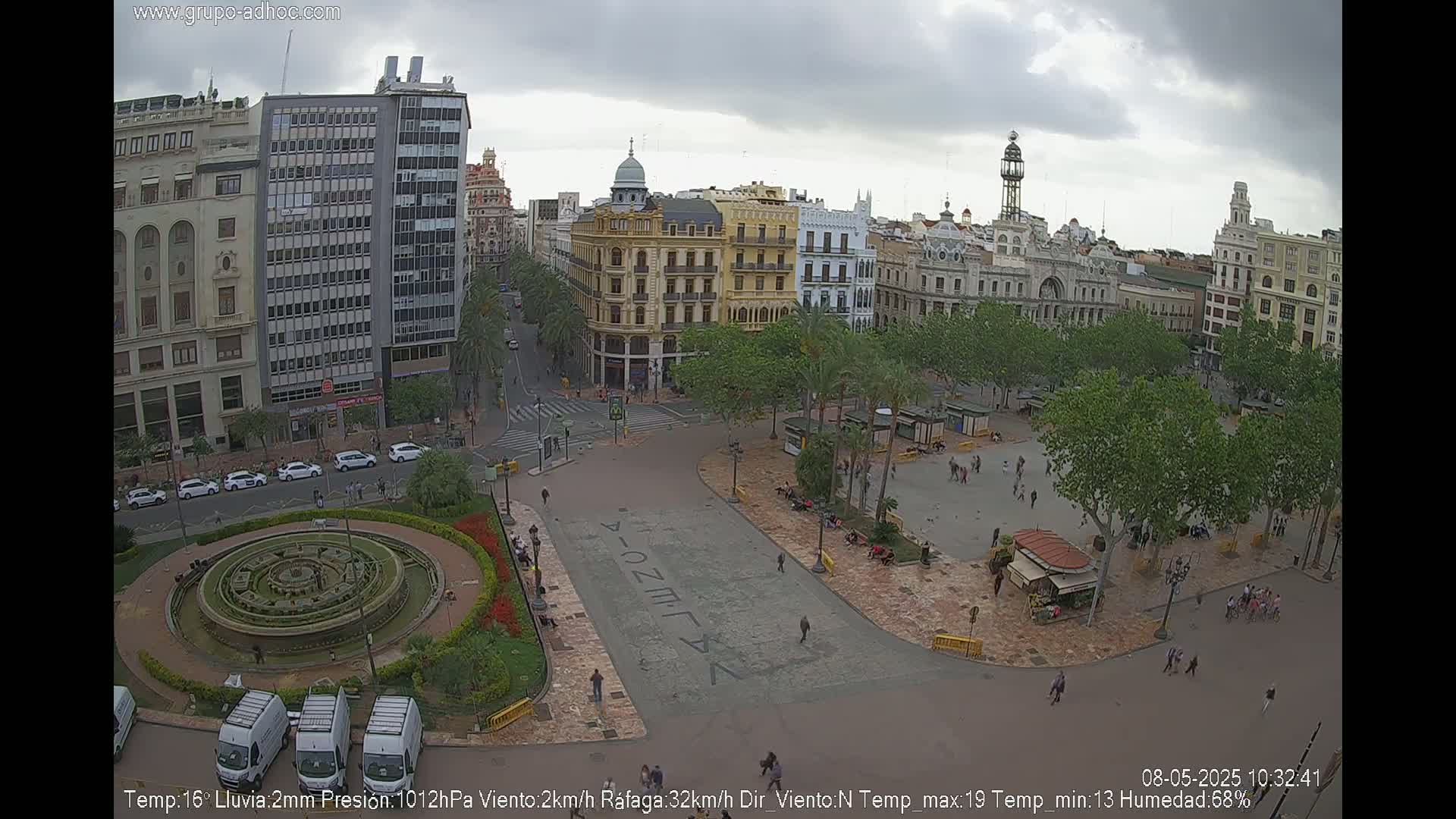 An aerial view of a city square with a fountain, surrounded by buildings under an overcast sky.