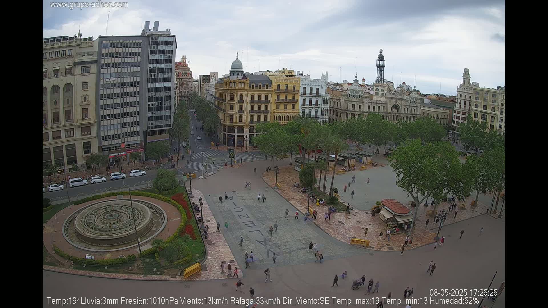 A city square with a fountain, surrounded by buildings and trees under an overcast sky.