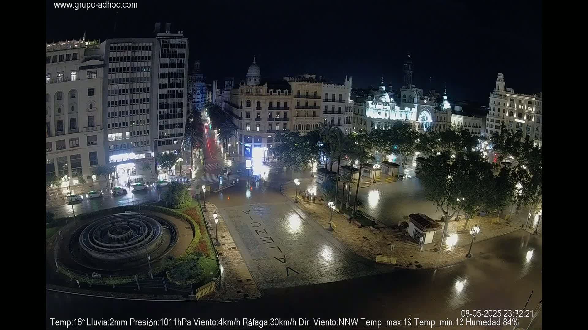 A nighttime aerial view of a city square partially flooded with rain, showing several buildings, a fountain, and palm trees lining the wet streets.