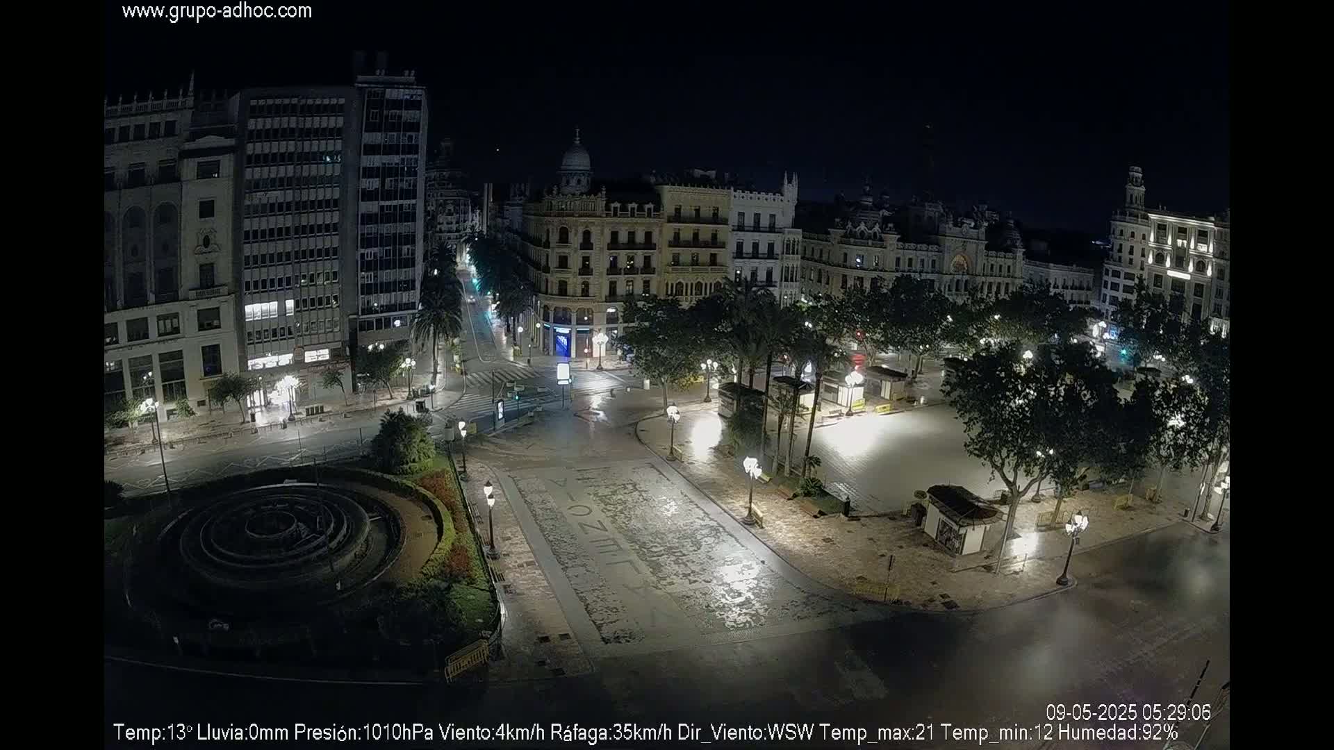 A nighttime aerial view of a city square with buildings surrounding it, illuminated by streetlights in wet conditions.