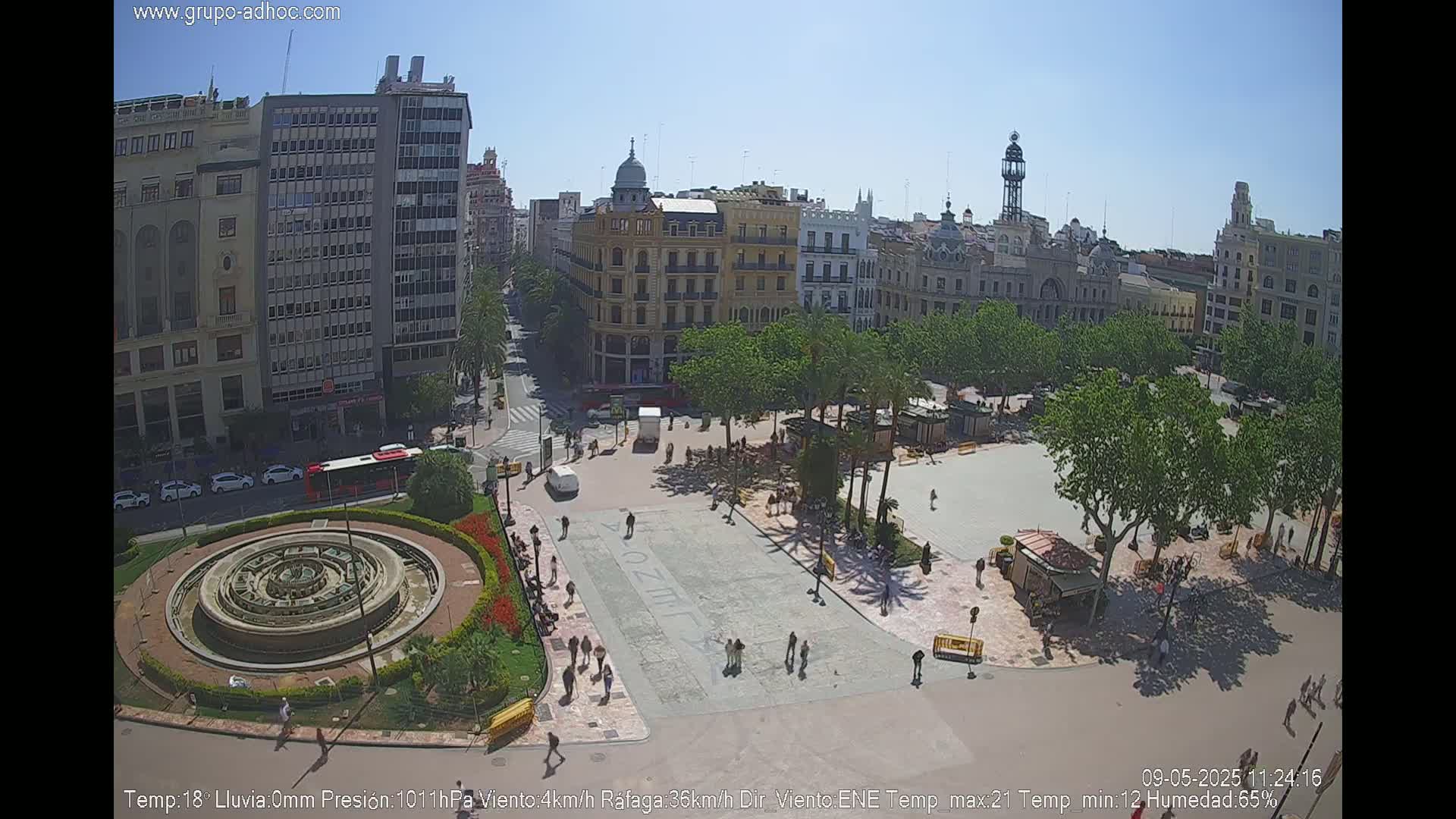 A sunny city square with a fountain, surrounded by buildings and palm trees, has people walking around.