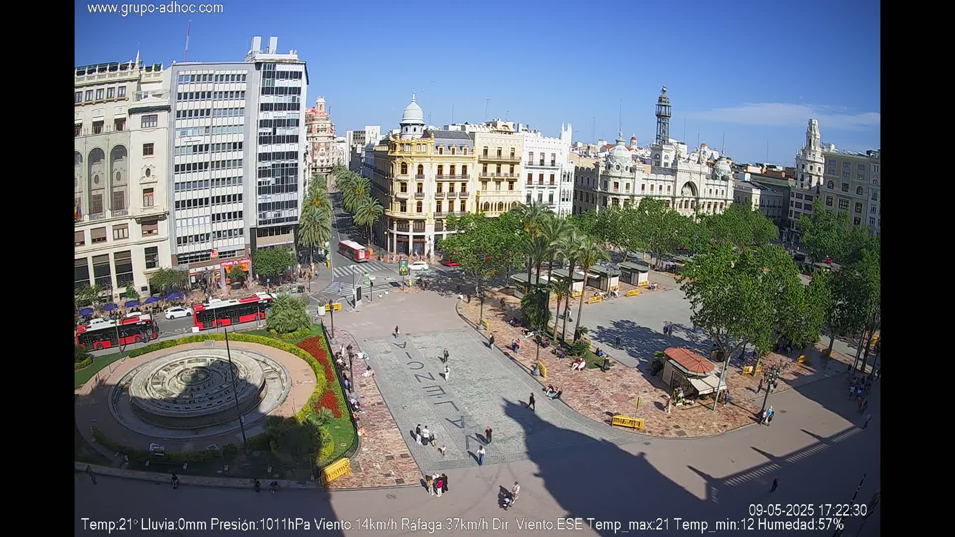 A sunny city square with a fountain, surrounded by buildings and palm trees, under a clear blue sky.
