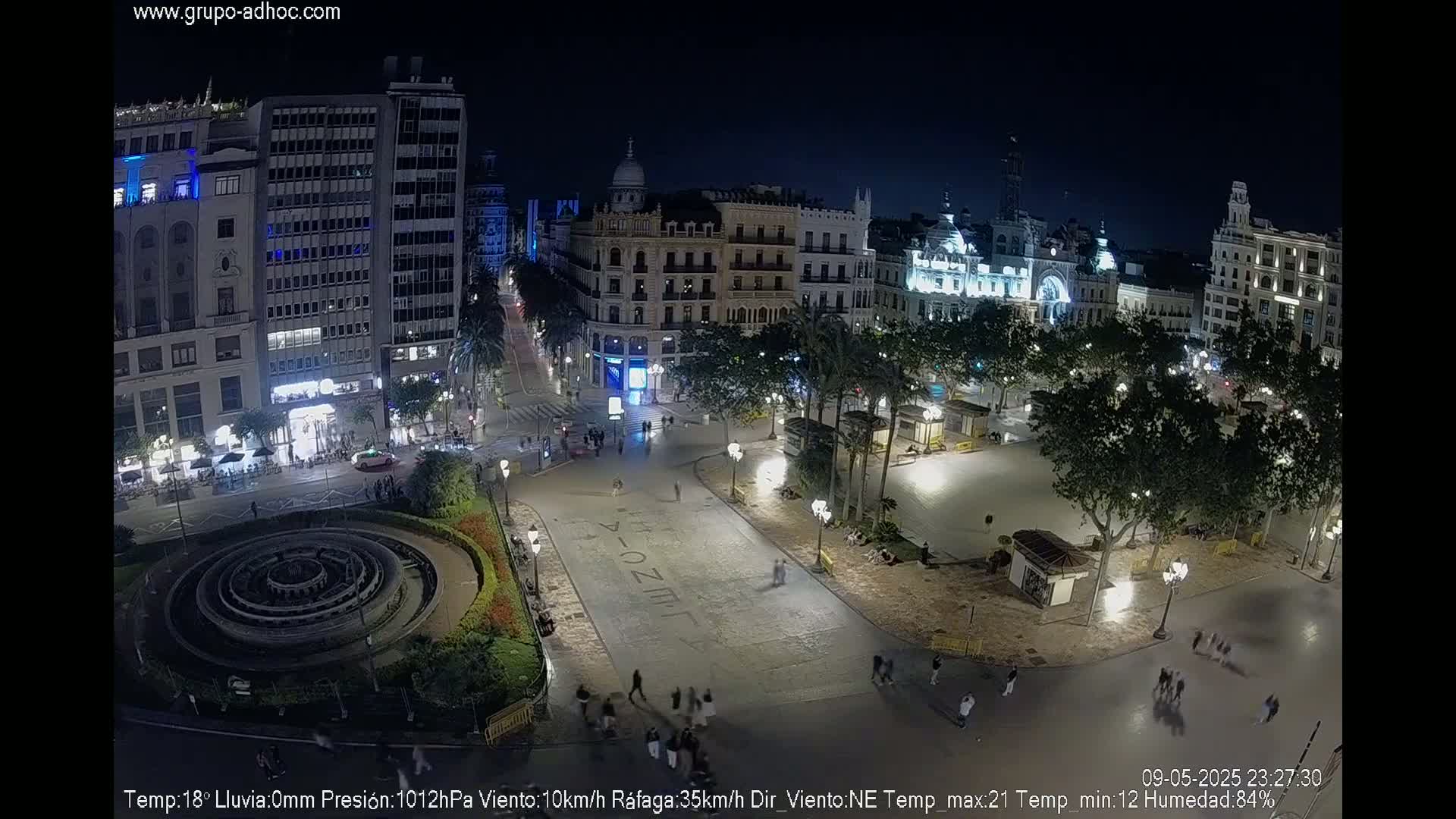 A nighttime aerial view of a city square with a circular fountain, surrounded by buildings and trees under a clear sky.