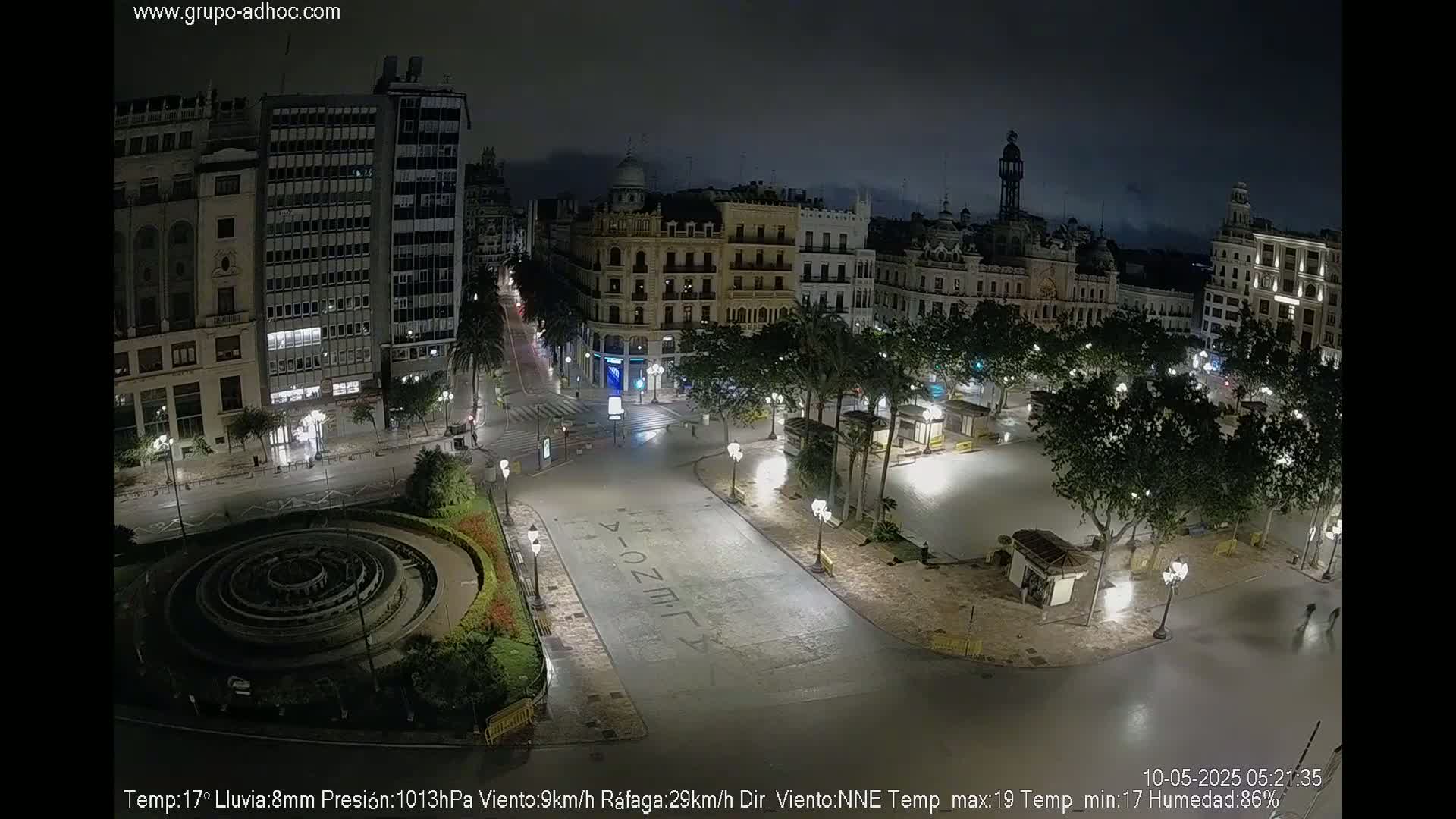 A nighttime view of a city square with a fountain, surrounded by buildings and trees under a rainy sky.