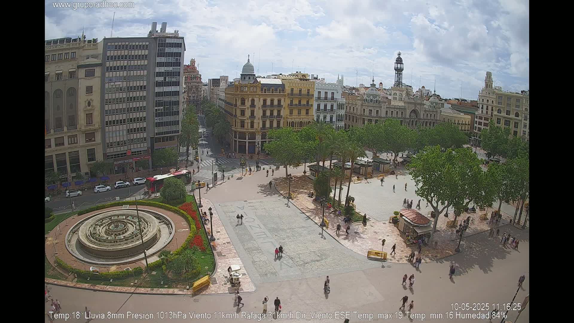 A city square with a fountain and surrounding buildings is shown on a partly cloudy day.