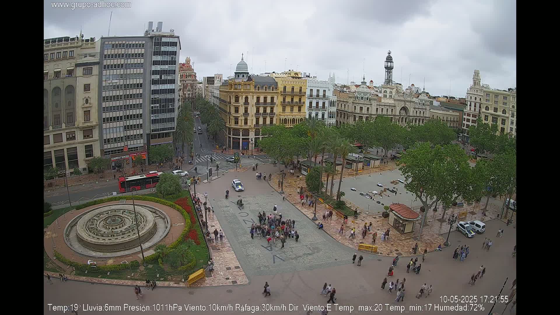 A city square with a fountain, surrounded by buildings and palm trees, under an overcast sky.