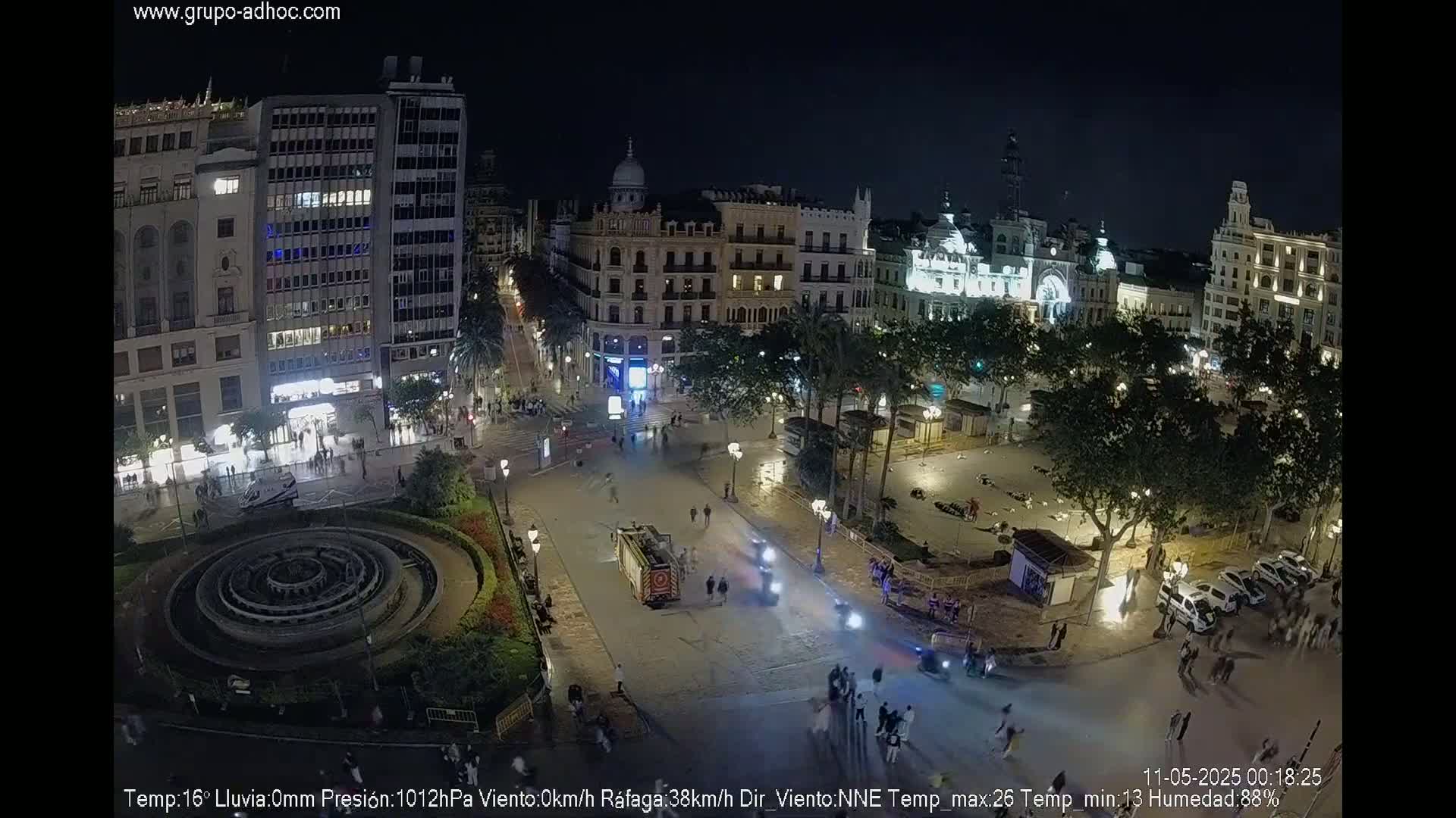 A nighttime aerial view of a city square with a circular fountain, surrounded by buildings and people, under a clear sky.