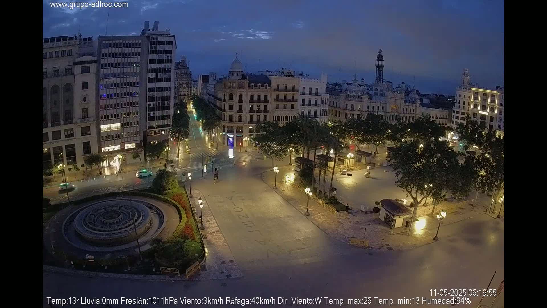 A nighttime view of a city square with a fountain, surrounded by buildings and palm trees under a dark, clear sky.