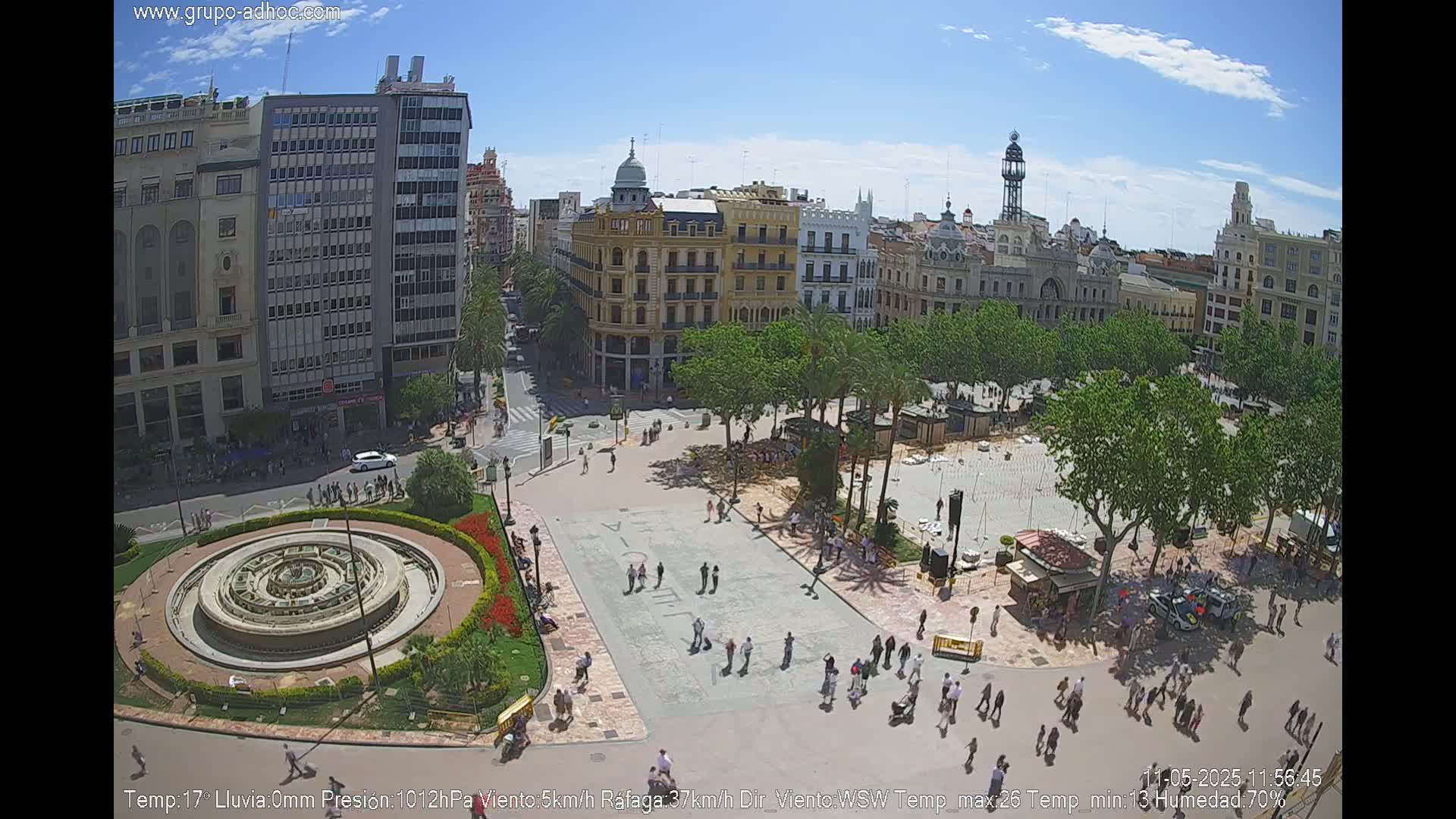 A sunny city square with a central fountain is surrounded by buildings and people walking under a clear blue sky.
