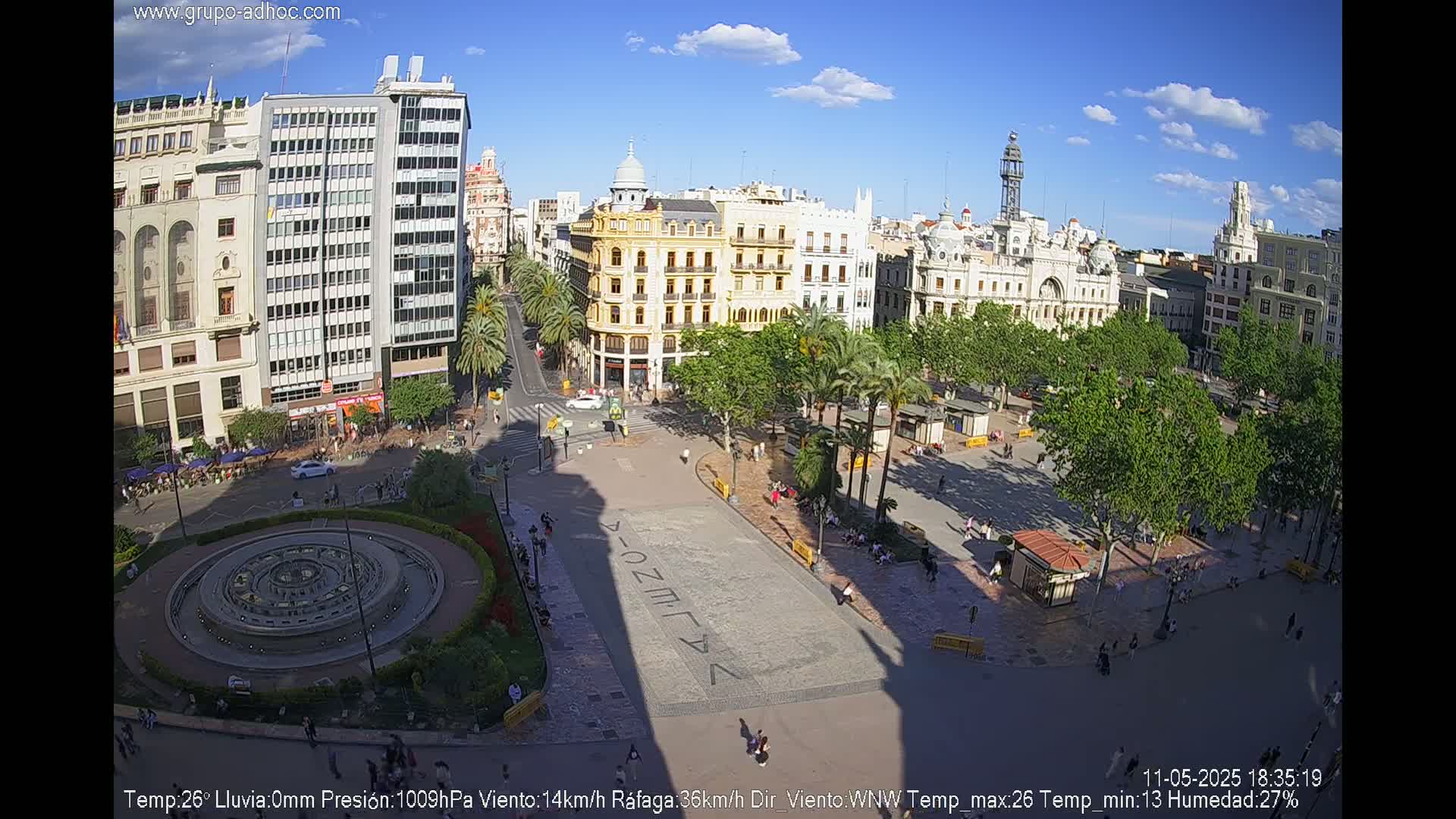 A sunny city square, filled with people and palm trees, is surrounded by tall buildings under a mostly clear sky.