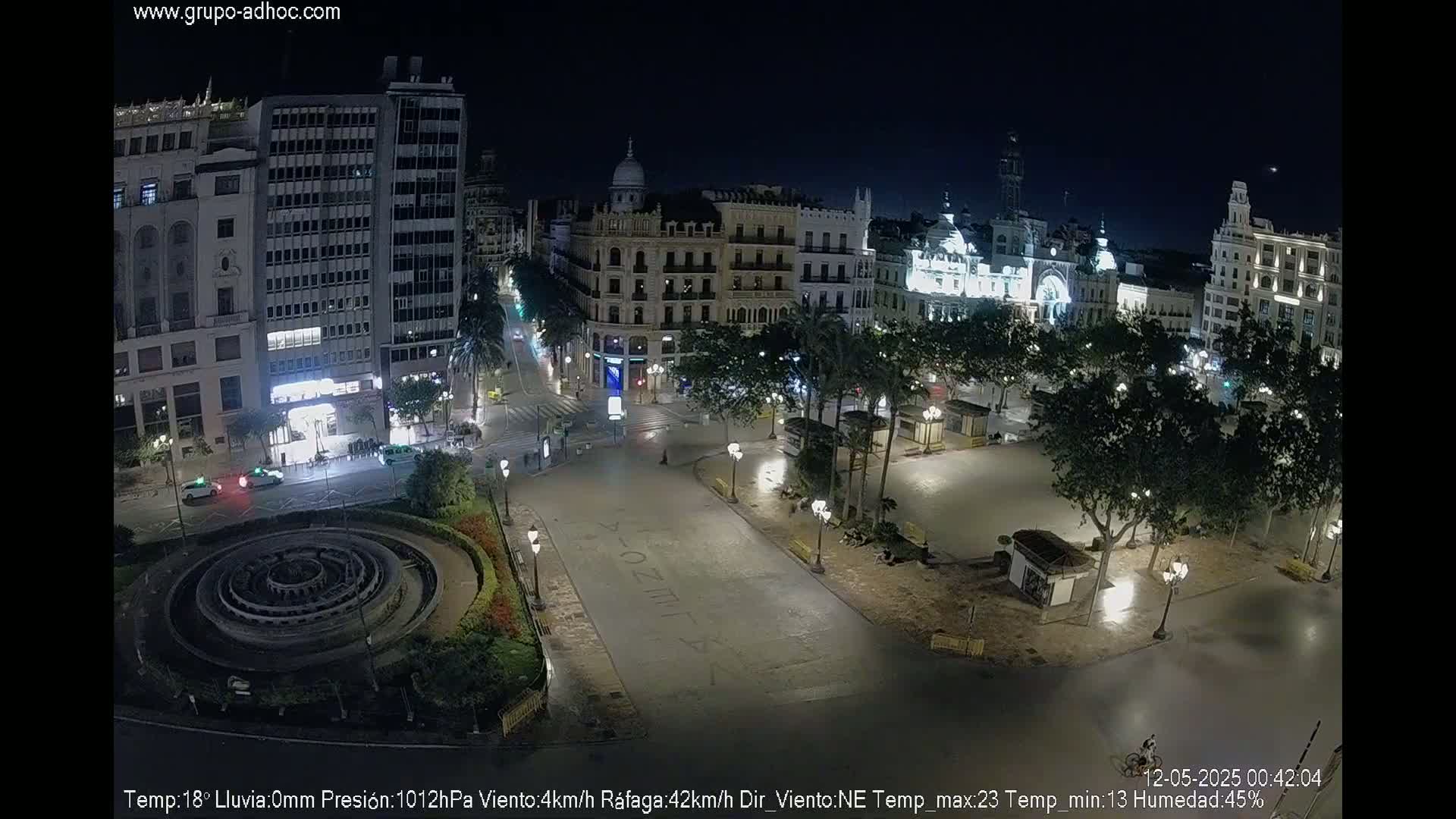 A nighttime, mostly empty city square with a circular fountain, surrounded by illuminated buildings and palm trees under a clear sky.