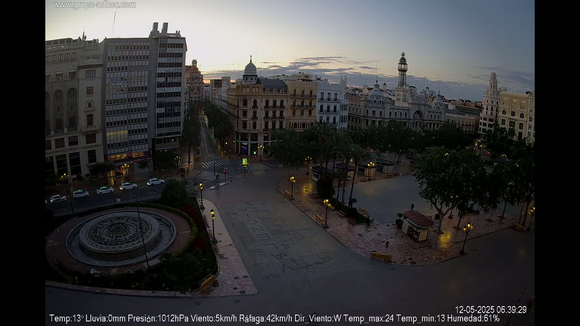 A mostly empty city square at dawn, with several buildings visible, a circular fountain in the foreground, and palm trees lining the square under a clear, dusky sky.
