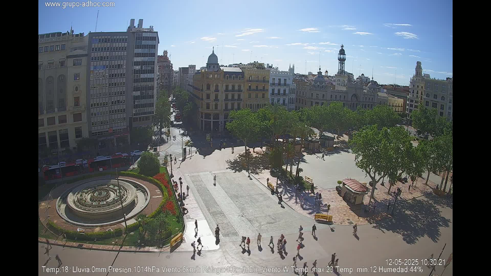 A sunny city square with a circular fountain, surrounded by buildings and trees, shows pedestrians walking on a paved plaza.
