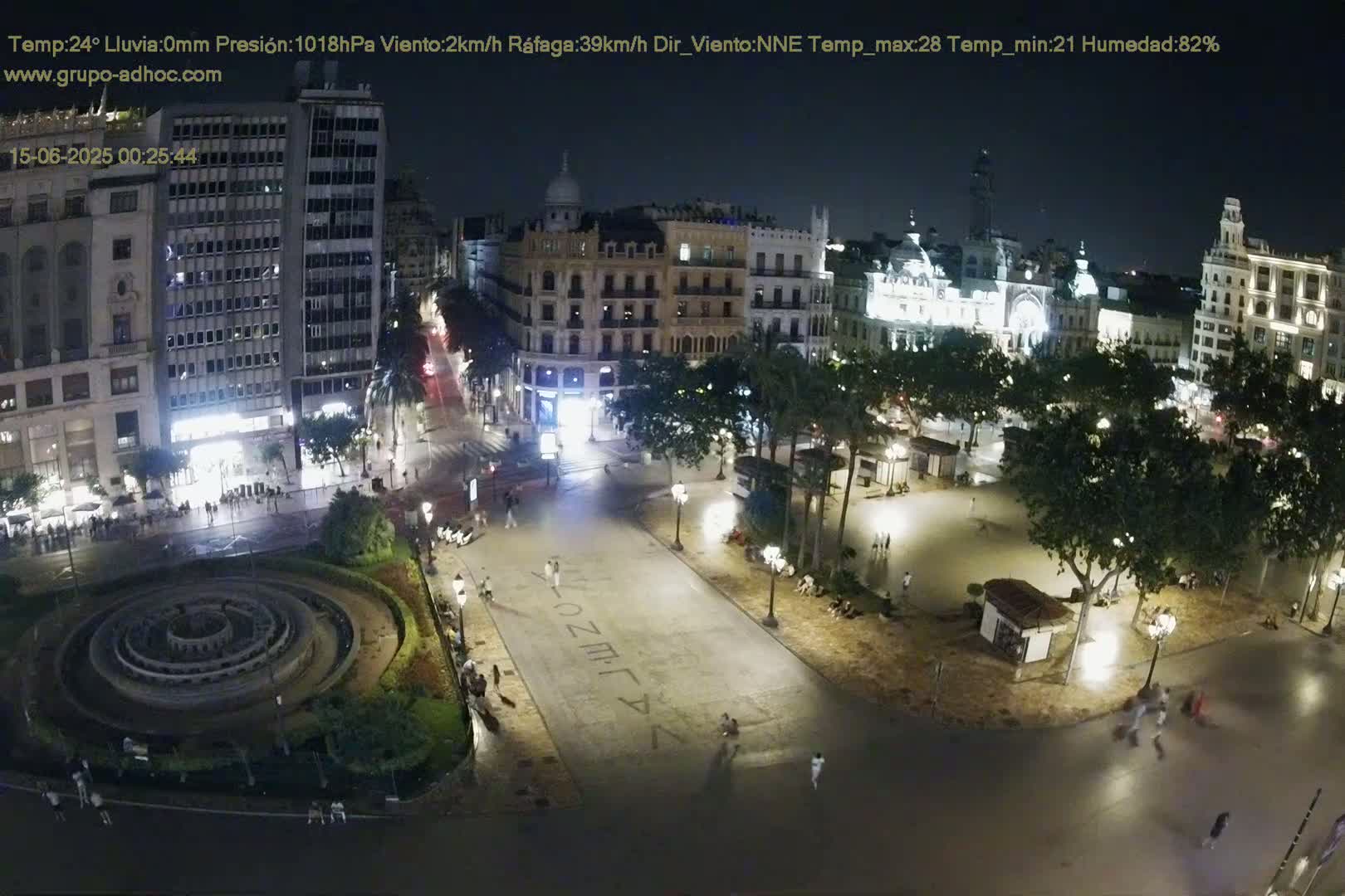 A sunny city square with a circular fountain, surrounded by buildings and trees, shows pedestrians walking on a paved plaza.