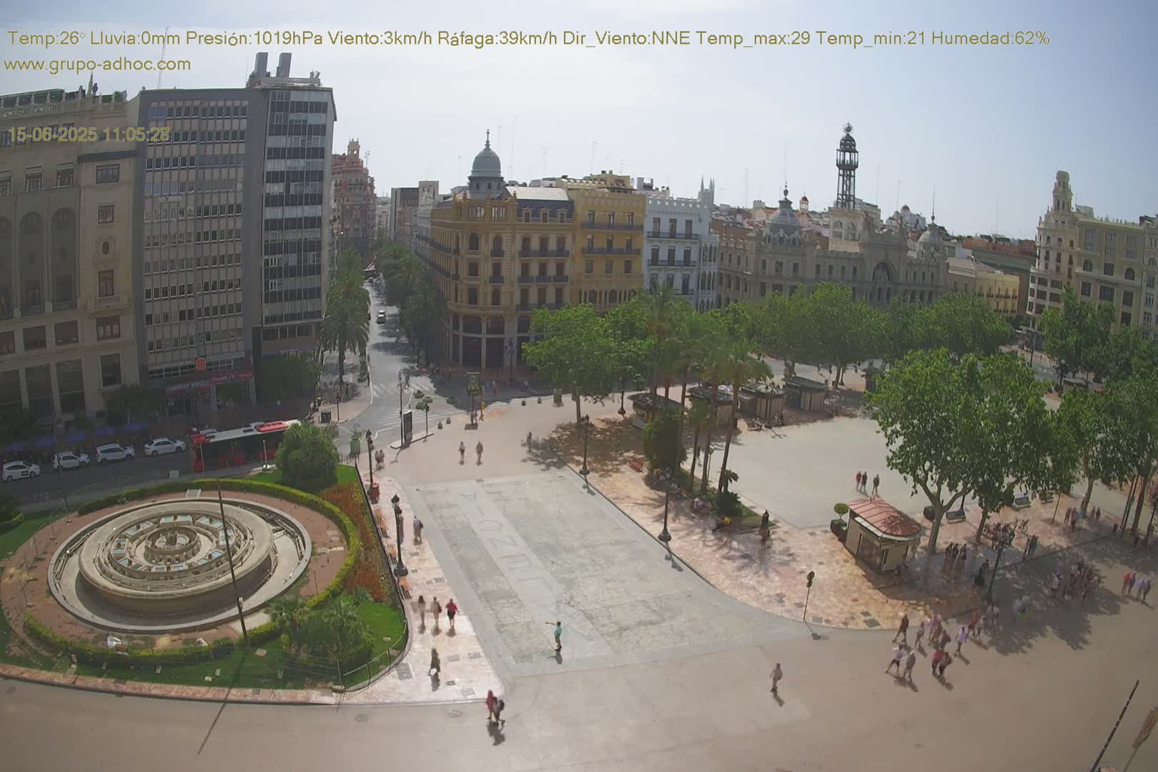 A sunny city square with a circular fountain, surrounded by buildings and trees, shows people walking around.