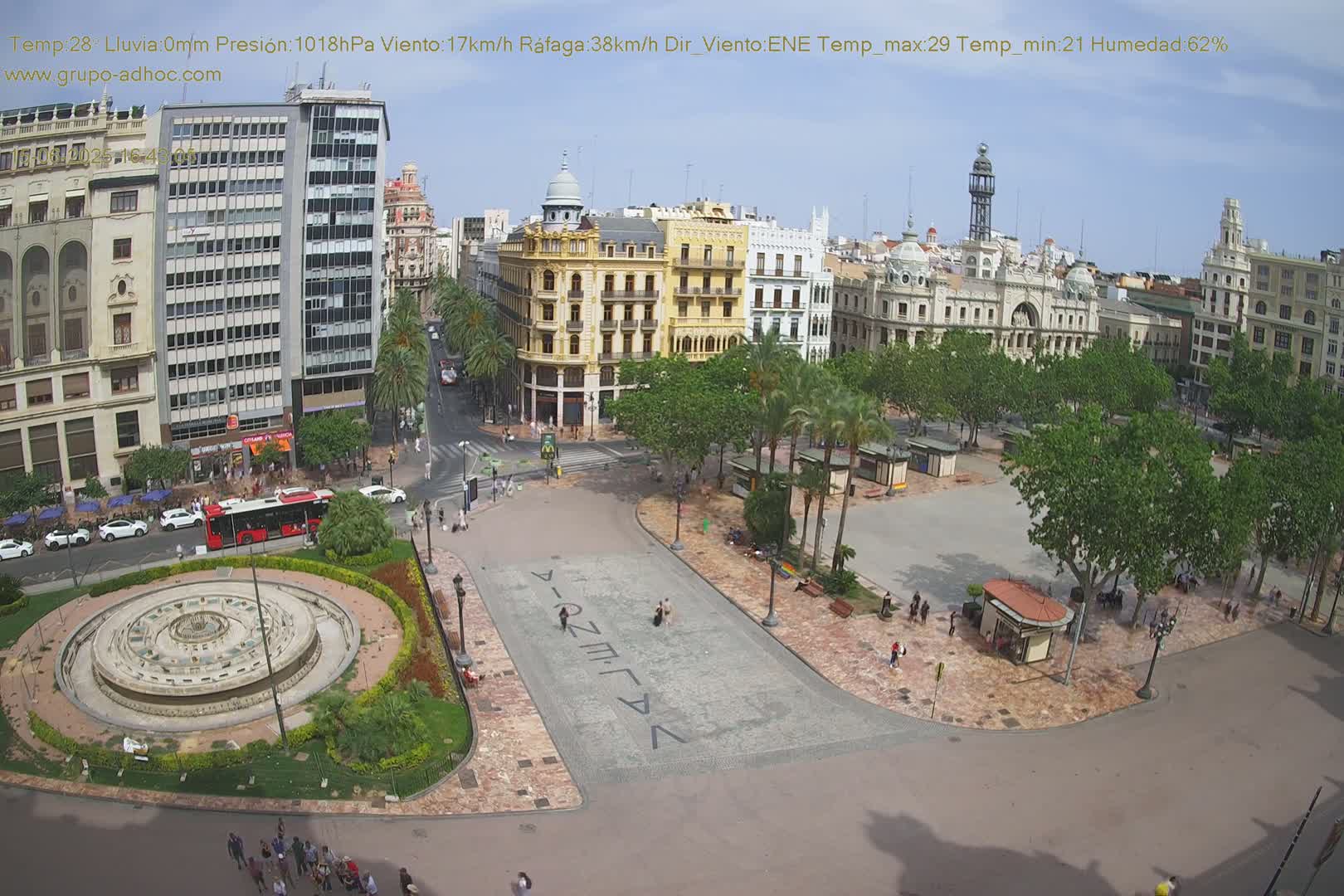 A high-angle view of a city square on a sunny day, showing a fountain, several buildings, palm trees, and people walking around.