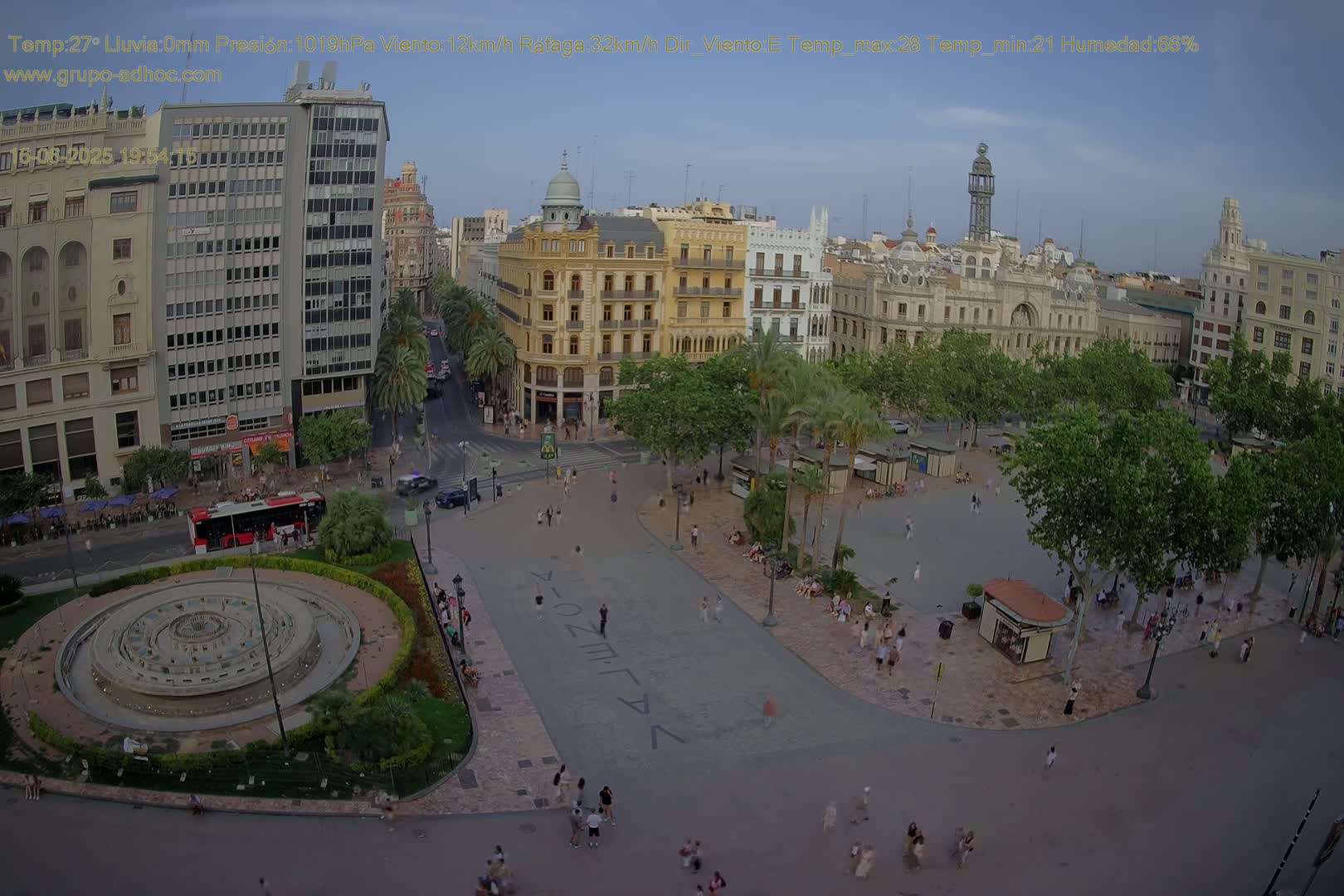 A city square with a fountain and many people, surrounded by buildings and trees, on a clear, sunny day.