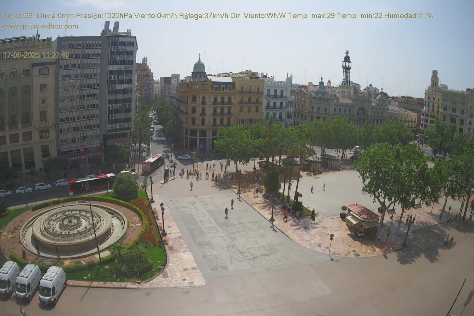 An aerial view of a city square on a sunny day, showing a circular fountain, several buildings, and people walking around.