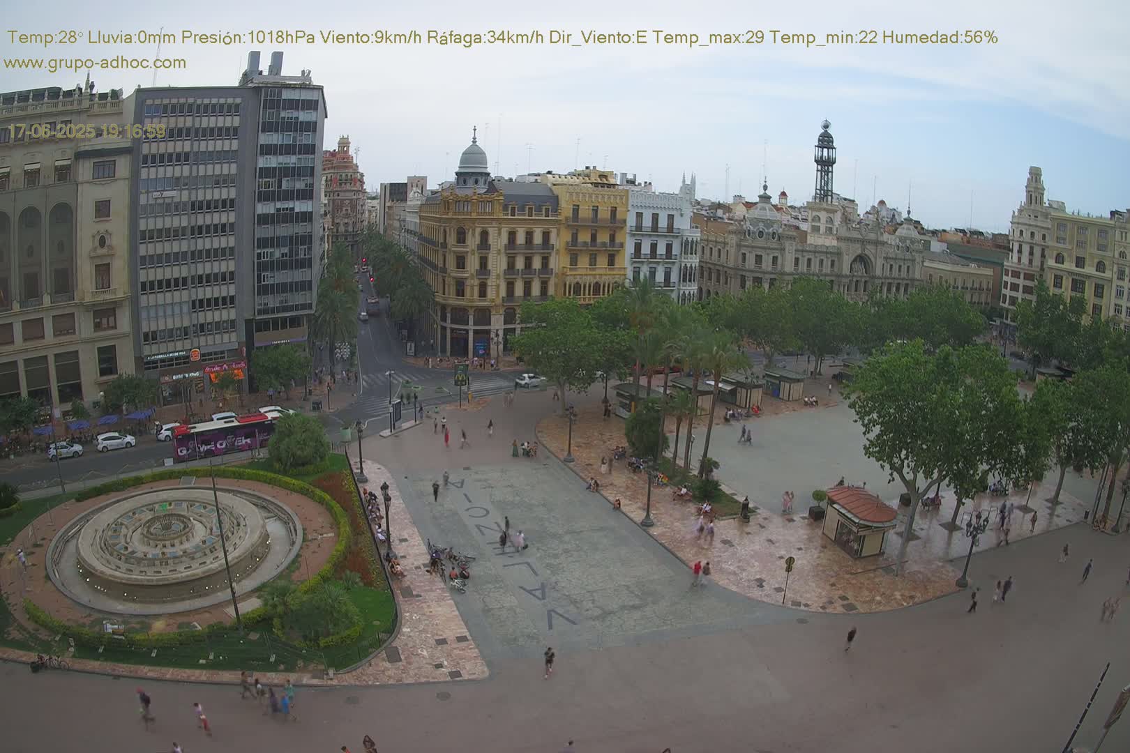 A city square with a central fountain, surrounded by buildings and palm trees, under a mostly cloudy sky.