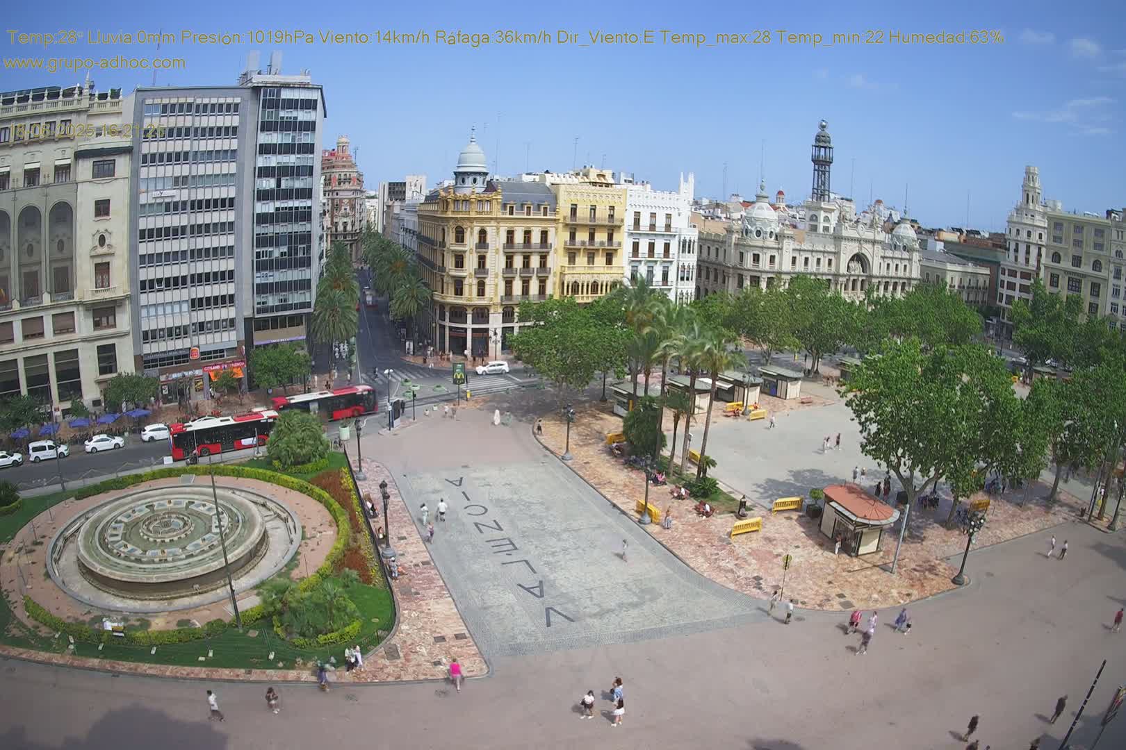 A city square with a fountain and several buildings surrounding it on a sunny day with a light breeze.