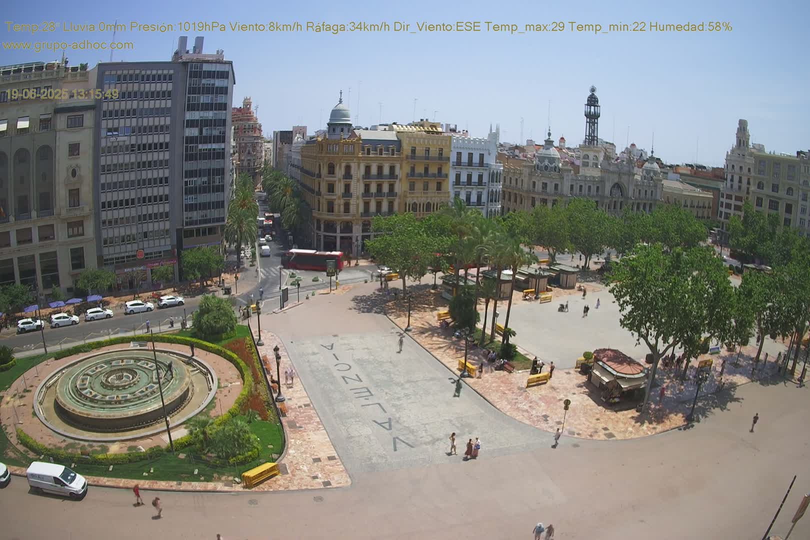 A sunny city square with a fountain, palm trees, and buildings, viewed from above.