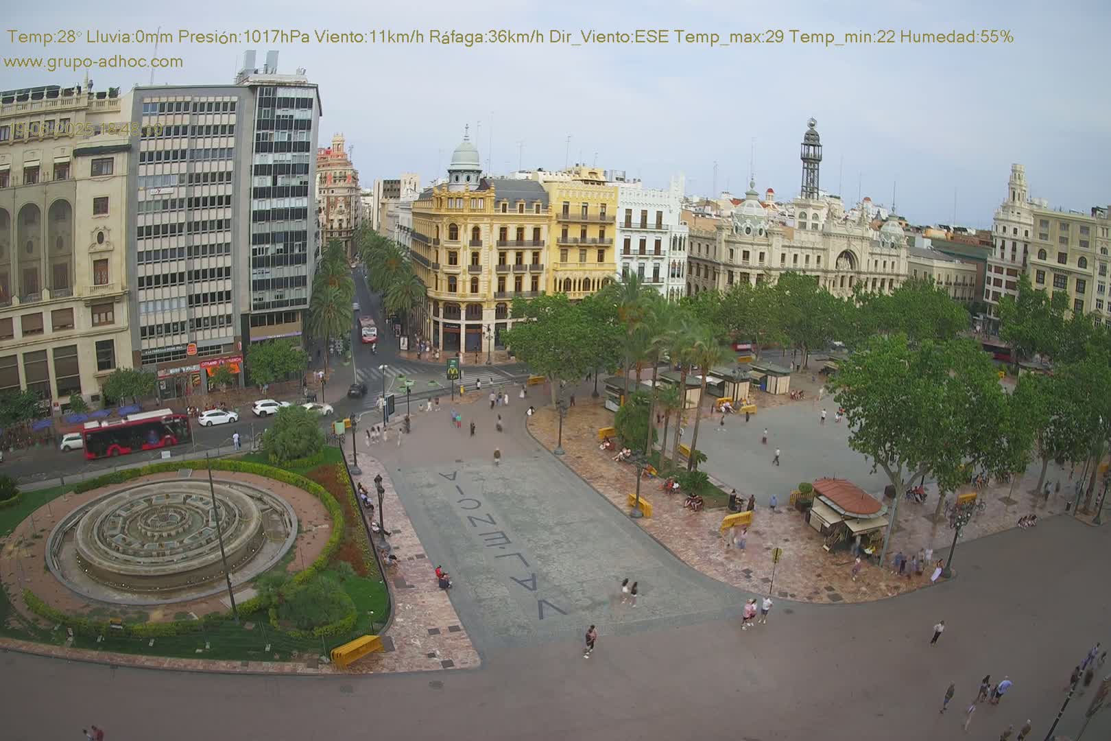 An aerial view of a city square with a central fountain, surrounded by buildings and trees under a mostly clear sky.