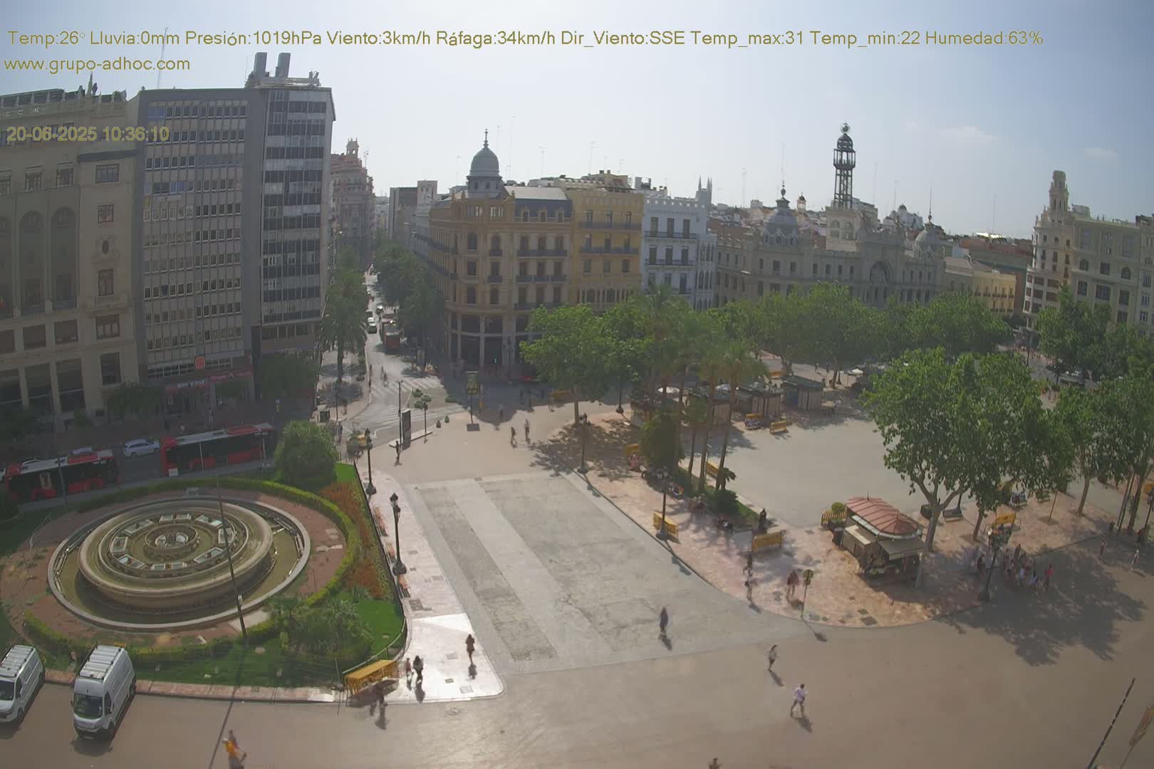 A sunny city square with a fountain, surrounded by buildings and trees, has people walking around.