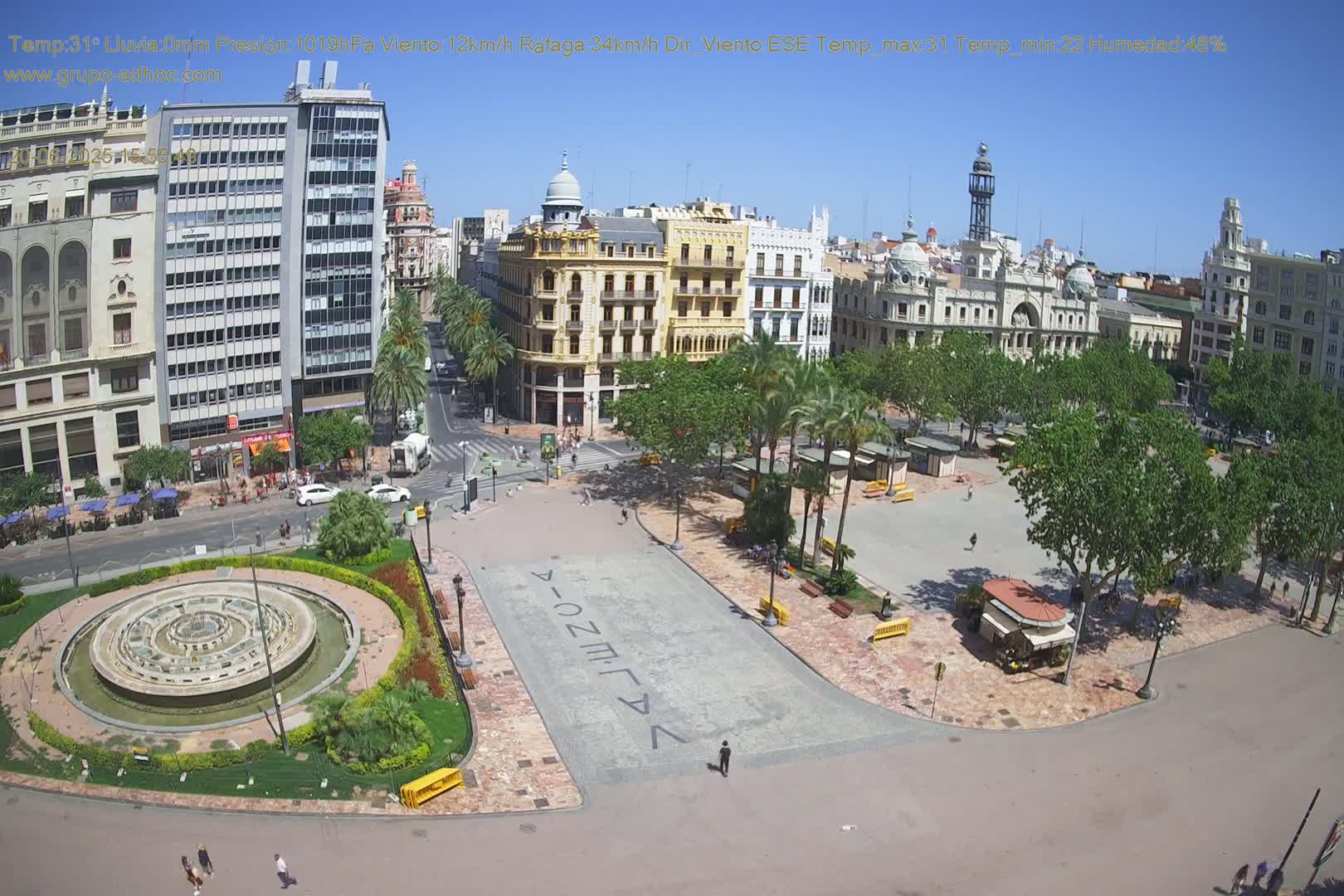 A city square with a circular fountain, palm trees, and surrounding buildings on a sunny day.