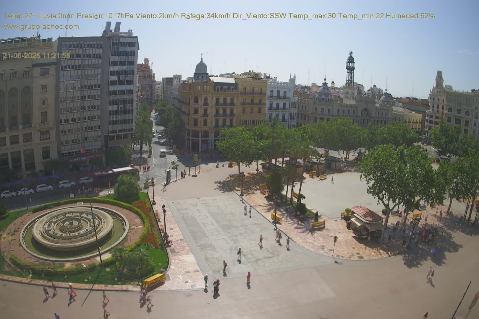 A sunny city square with a central fountain is surrounded by buildings and palm trees, with people walking around.