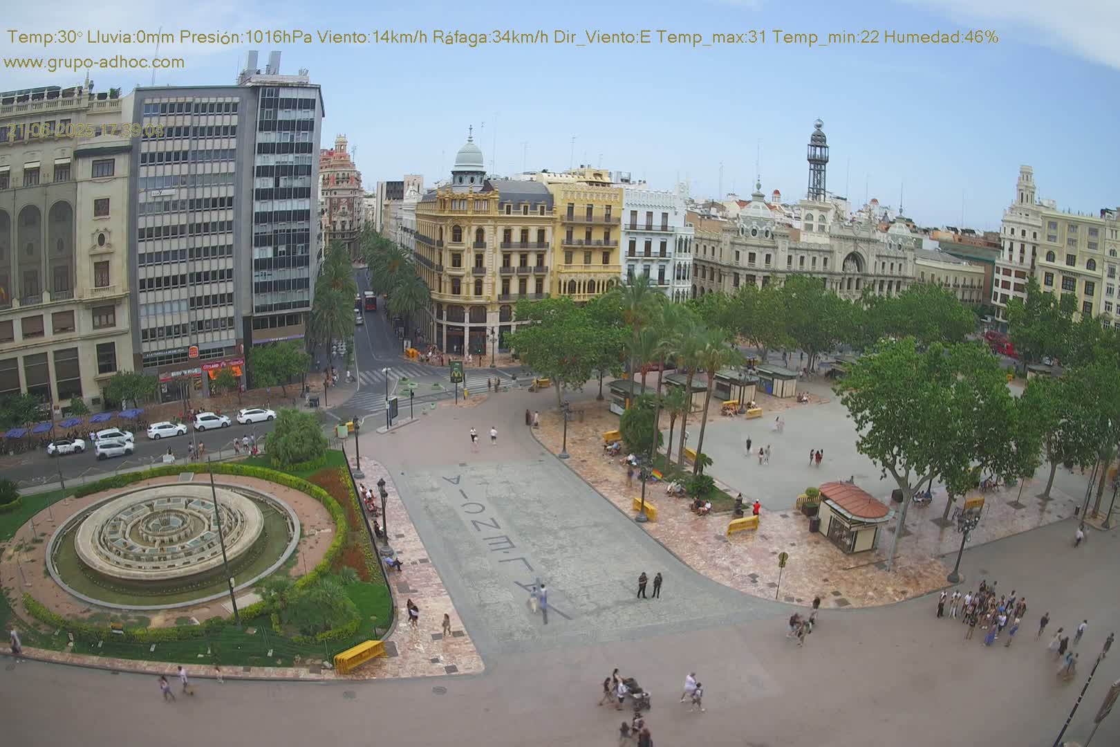A high-angle view of a city square with a central fountain, surrounded by buildings and palm trees under a clear, sunny sky.