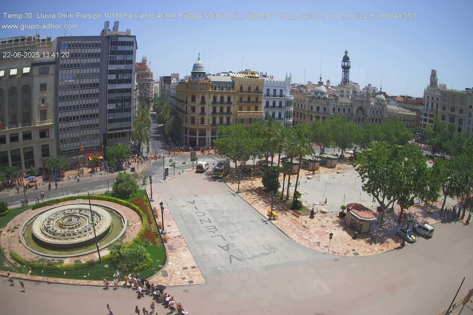 A sunny city square with a circular fountain, surrounded by buildings and palm trees, under a clear blue sky.