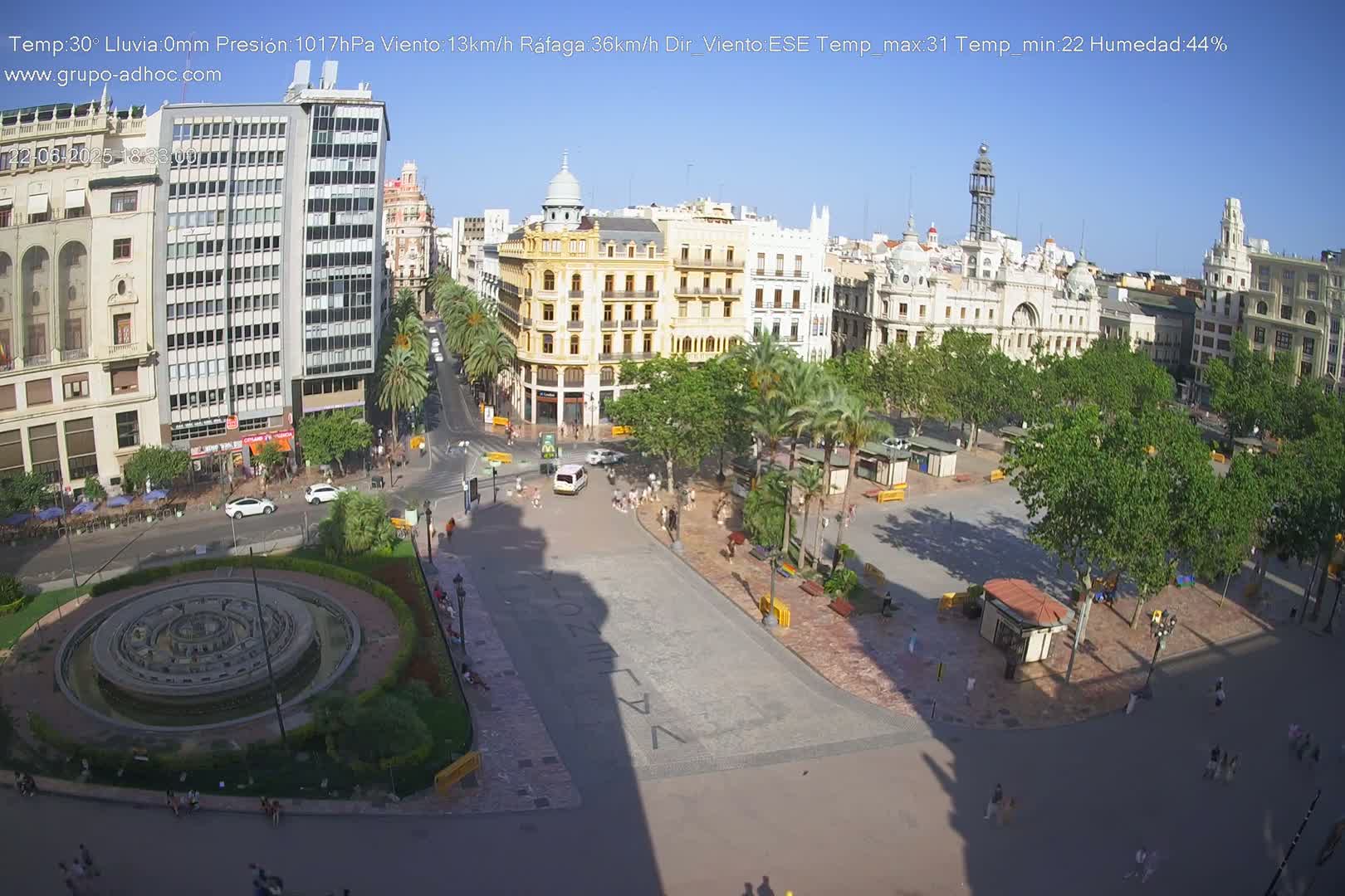 A sunny city square with a fountain, palm trees, and buildings surrounding it, under a clear blue sky.