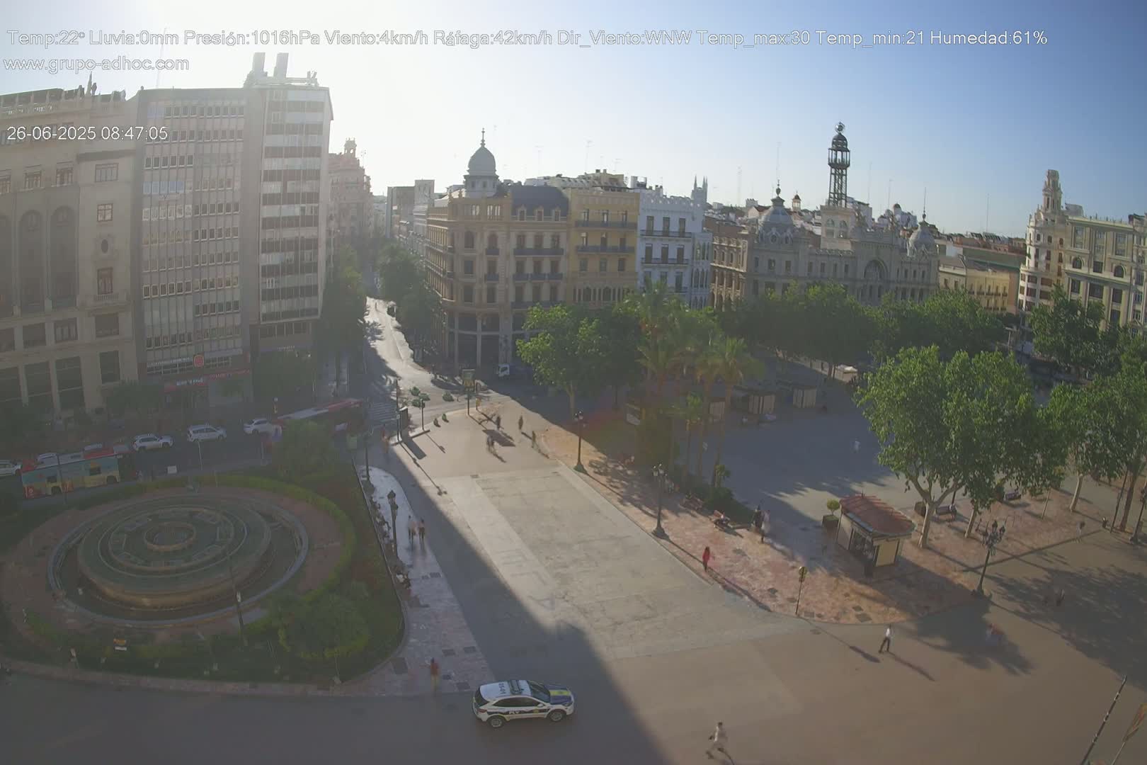 A sunny city square with a circular fountain, surrounded by buildings and trees, with a police car parked nearby.
