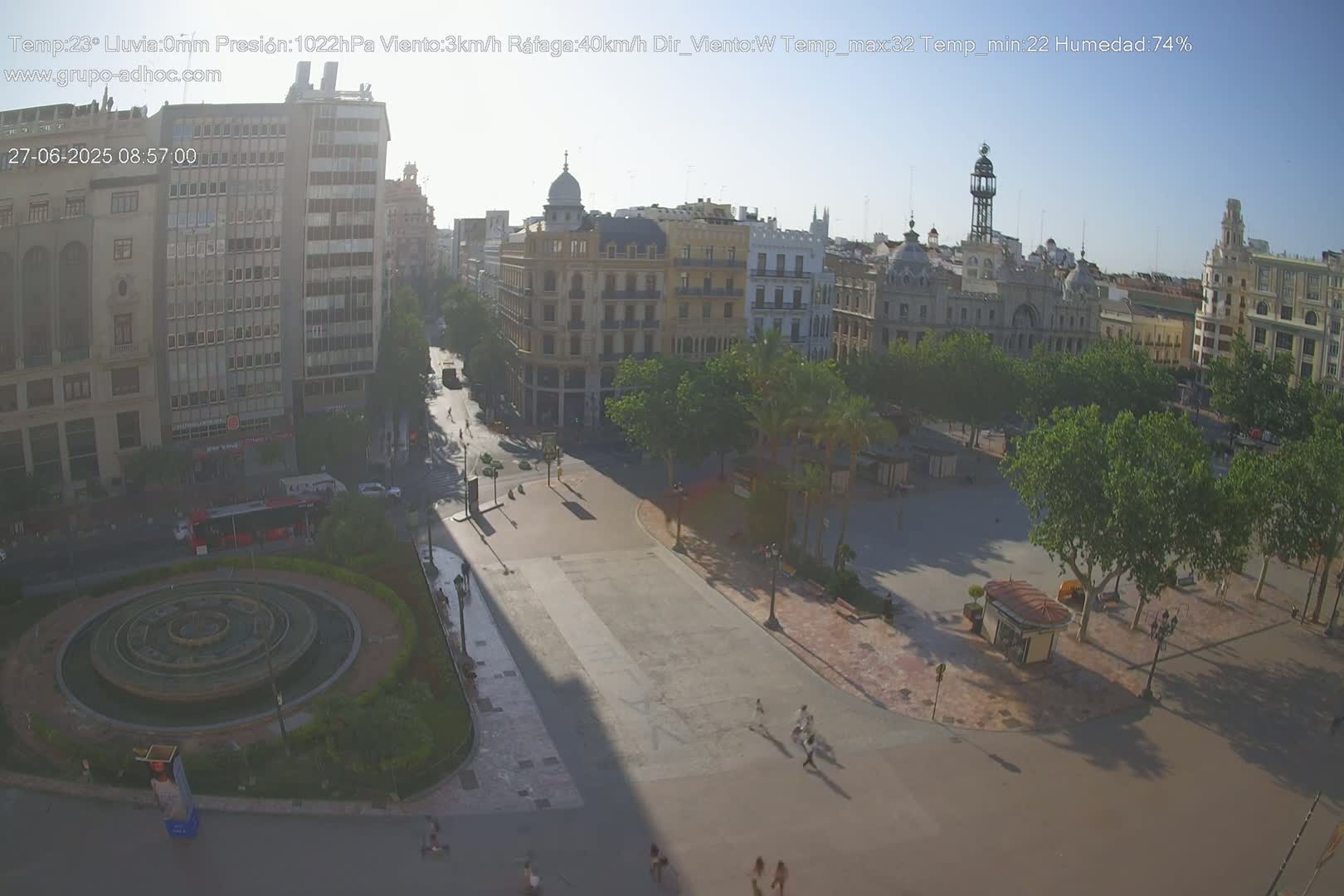A mostly empty city square with a circular fountain, surrounded by buildings and trees, on a sunny day with light haze.
