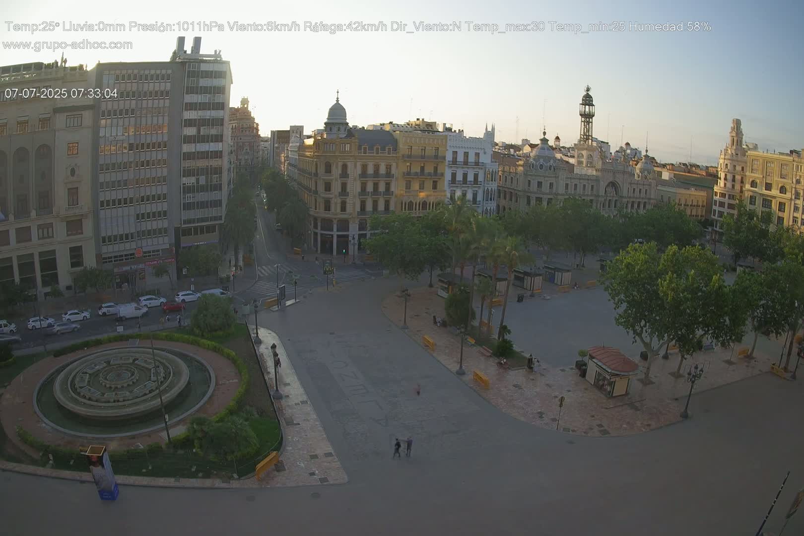 A city square with a fountain, surrounded by buildings and trees, under a clear, bright sky.