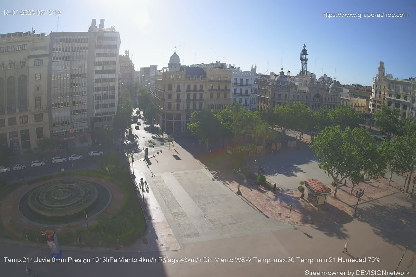 A sunny city square with a fountain and surrounding buildings is viewed from above, with some trees providing shade.