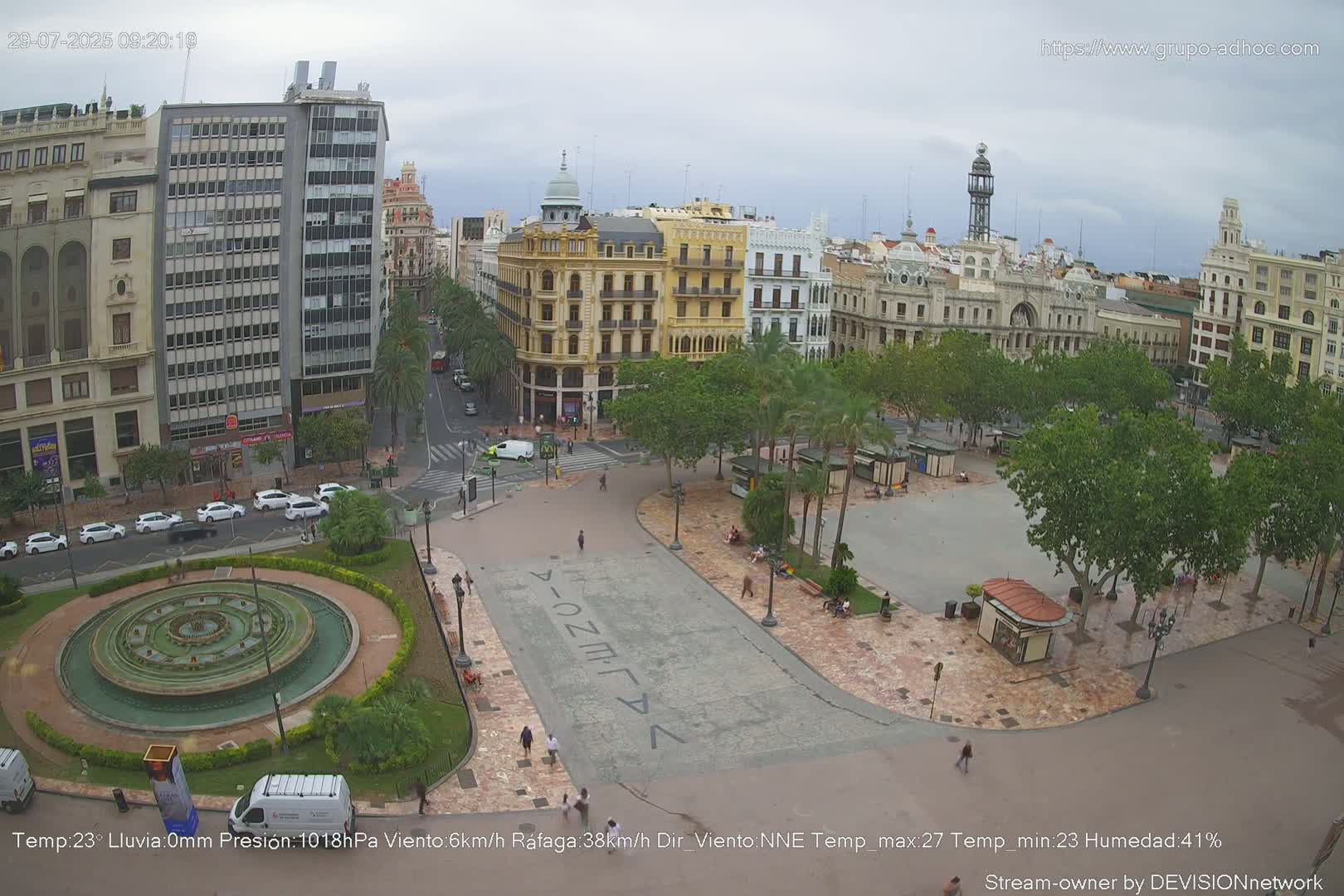 A city square with a fountain, palm trees, and buildings surrounding it under an overcast sky.