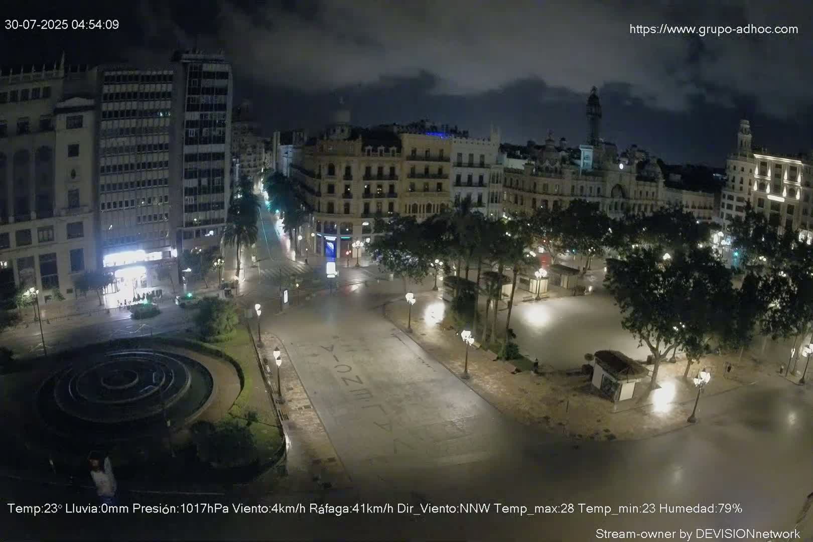A city square with a fountain, palm trees, and buildings surrounding it under an overcast sky.