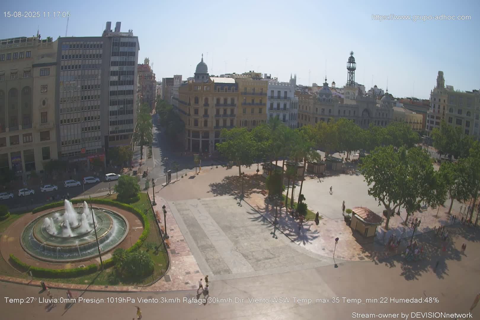 A city square with a fountain, palm trees, and buildings surrounding it under an overcast sky.