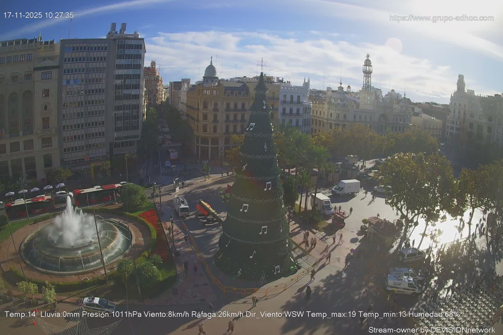 A vibrant city plaza, adorned with a large Christmas tree decorated with musical notes and a central fountain, is bustling with people and vehicles under a clear, sunny sky with scattered clouds.