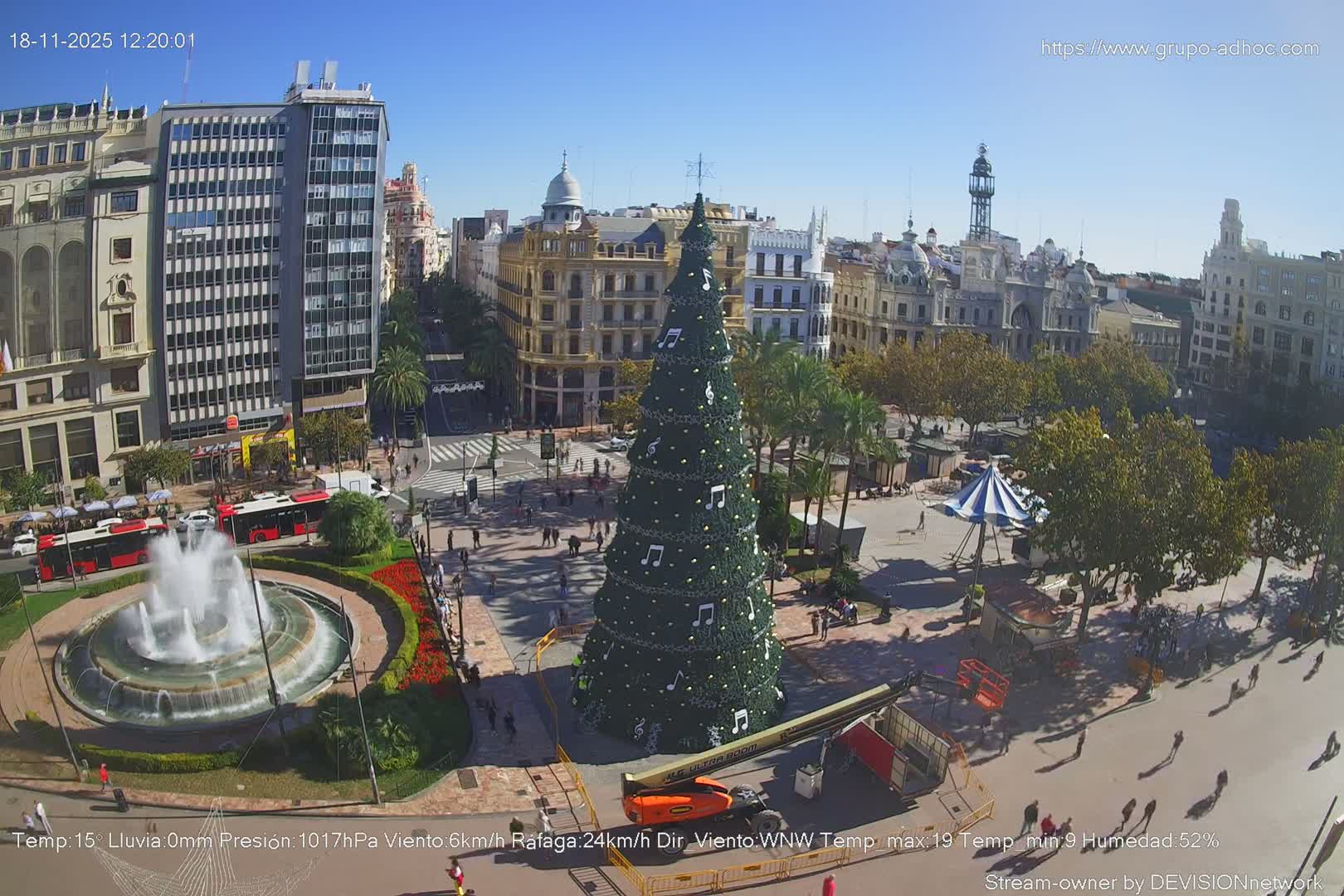 A vibrant city plaza, adorned with a large Christmas tree decorated with musical notes and a central fountain, is bustling with people and vehicles under a clear, sunny sky with scattered clouds.