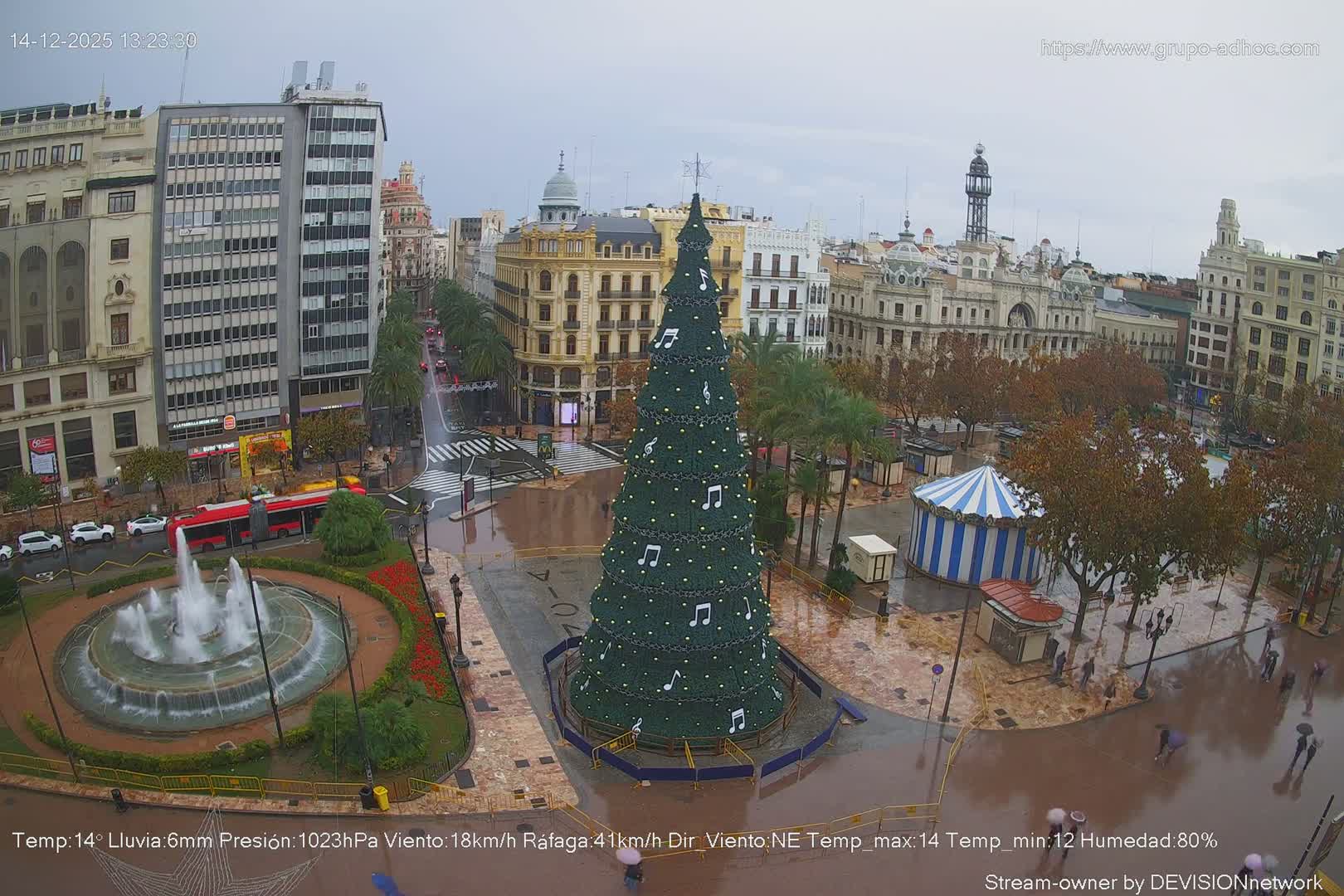 A vibrant city plaza, adorned with a large Christmas tree decorated with musical notes and a central fountain, is bustling with people and vehicles under a clear, sunny sky with scattered clouds.
