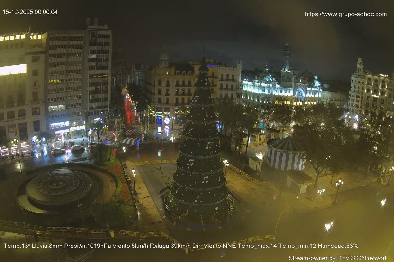 A vibrant city plaza, adorned with a large Christmas tree decorated with musical notes and a central fountain, is bustling with people and vehicles under a clear, sunny sky with scattered clouds.