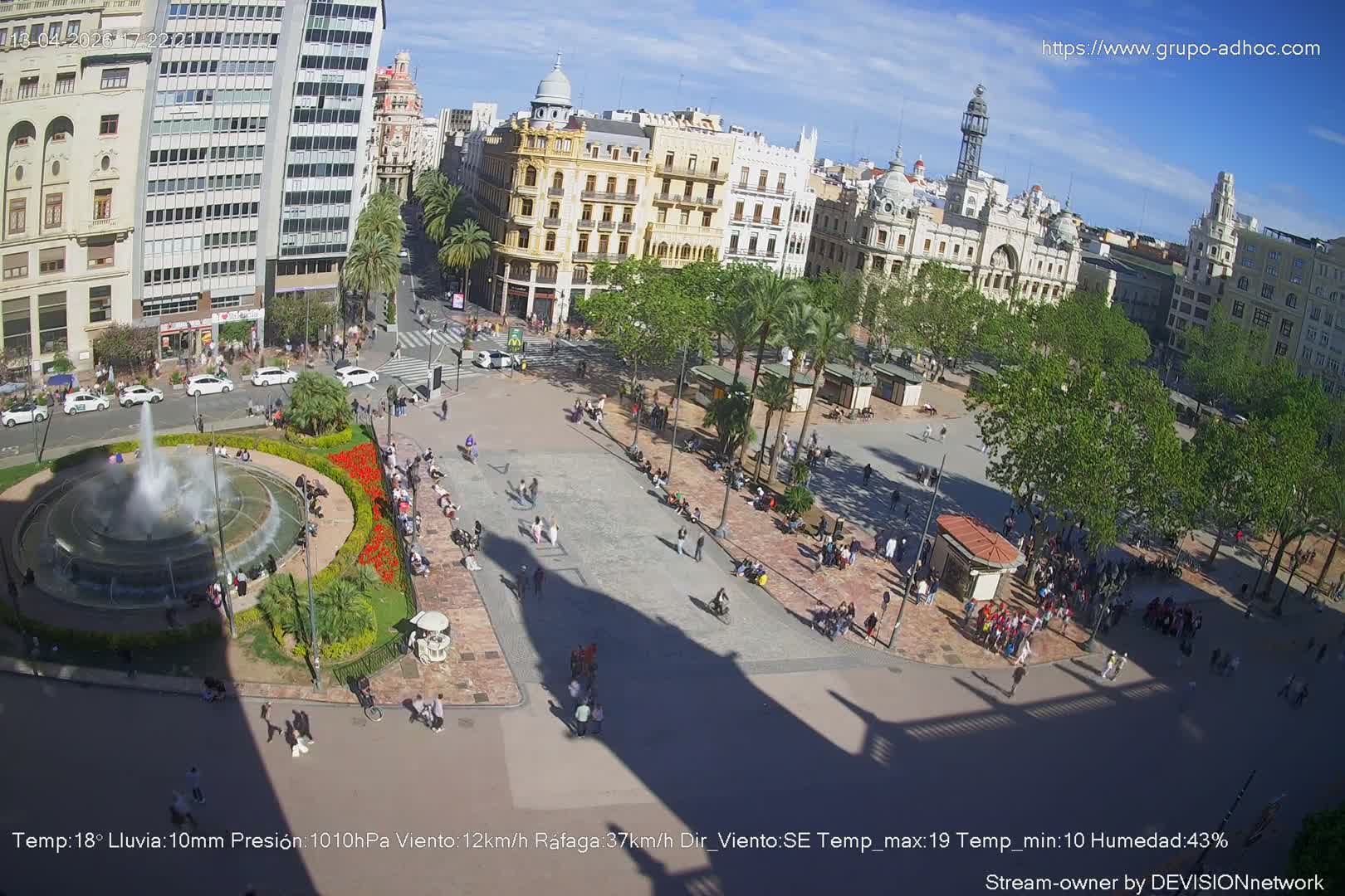 A vibrant city plaza, adorned with a large Christmas tree decorated with musical notes and a central fountain, is bustling with people and vehicles under a clear, sunny sky with scattered clouds.