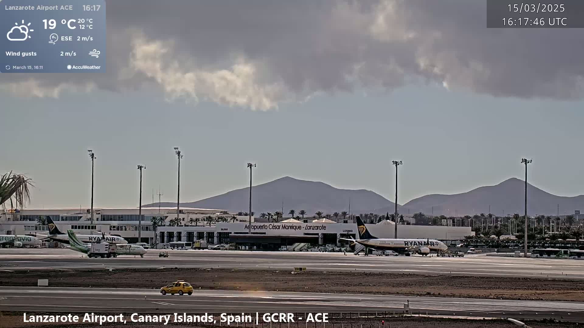 A partly cloudy daytime scene at an airport shows several airplanes on the tarmac, with mountains visible in the distance.