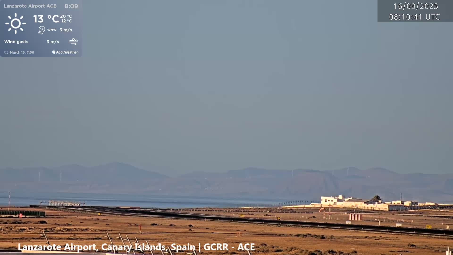 A hazy, sunny day at an airport shows a runway, some buildings, and a body of water with distant mountains in the background.