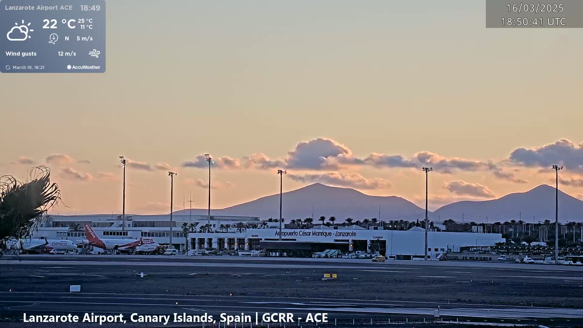 An airport tarmac with several airplanes and airport buildings is visible under a partly cloudy sunset sky with a mountain range in the background.