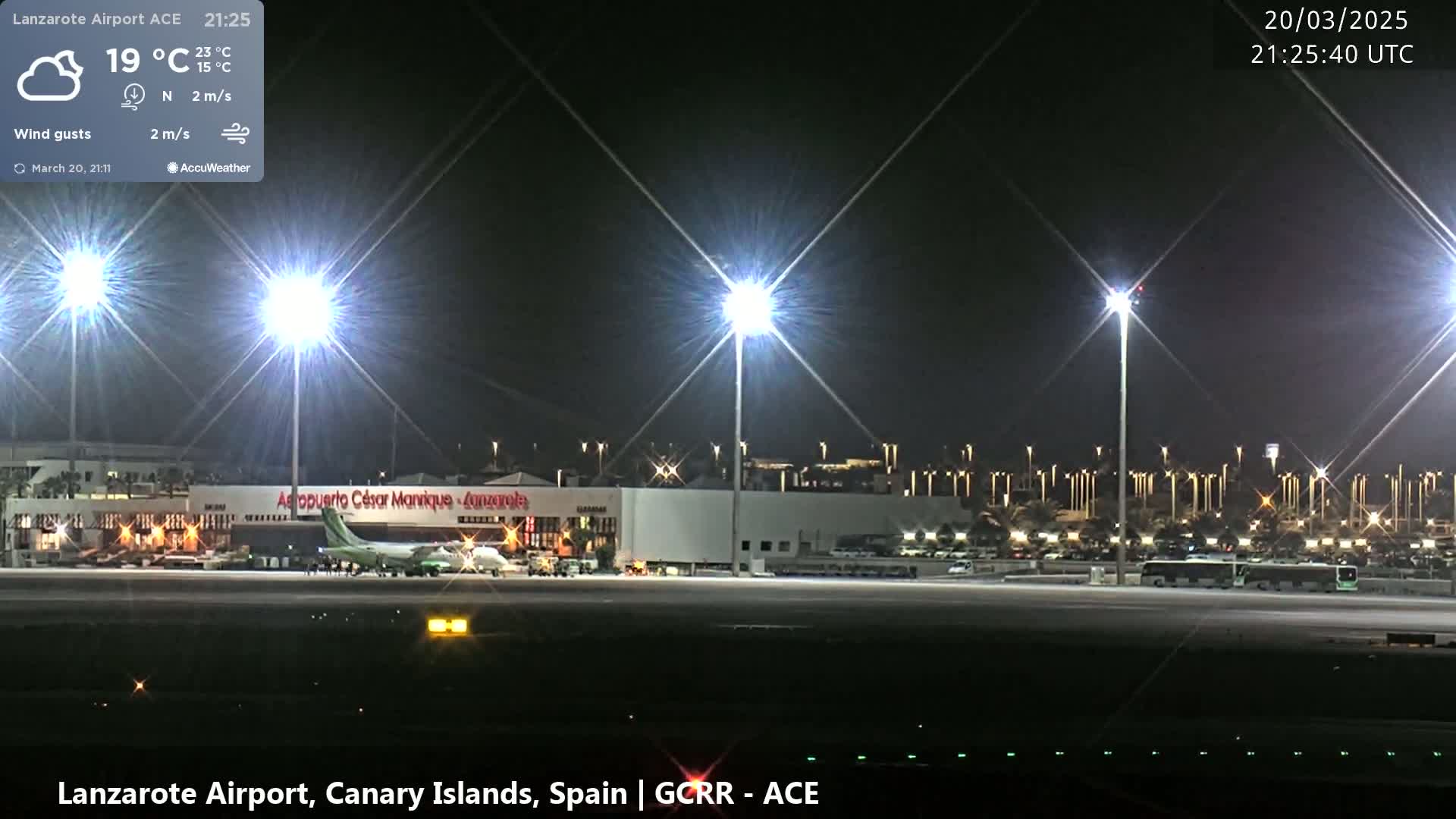 A nighttime view of an airport tarmac with a plane parked near a terminal building, brightly lit by numerous tall lights under a partly cloudy sky.