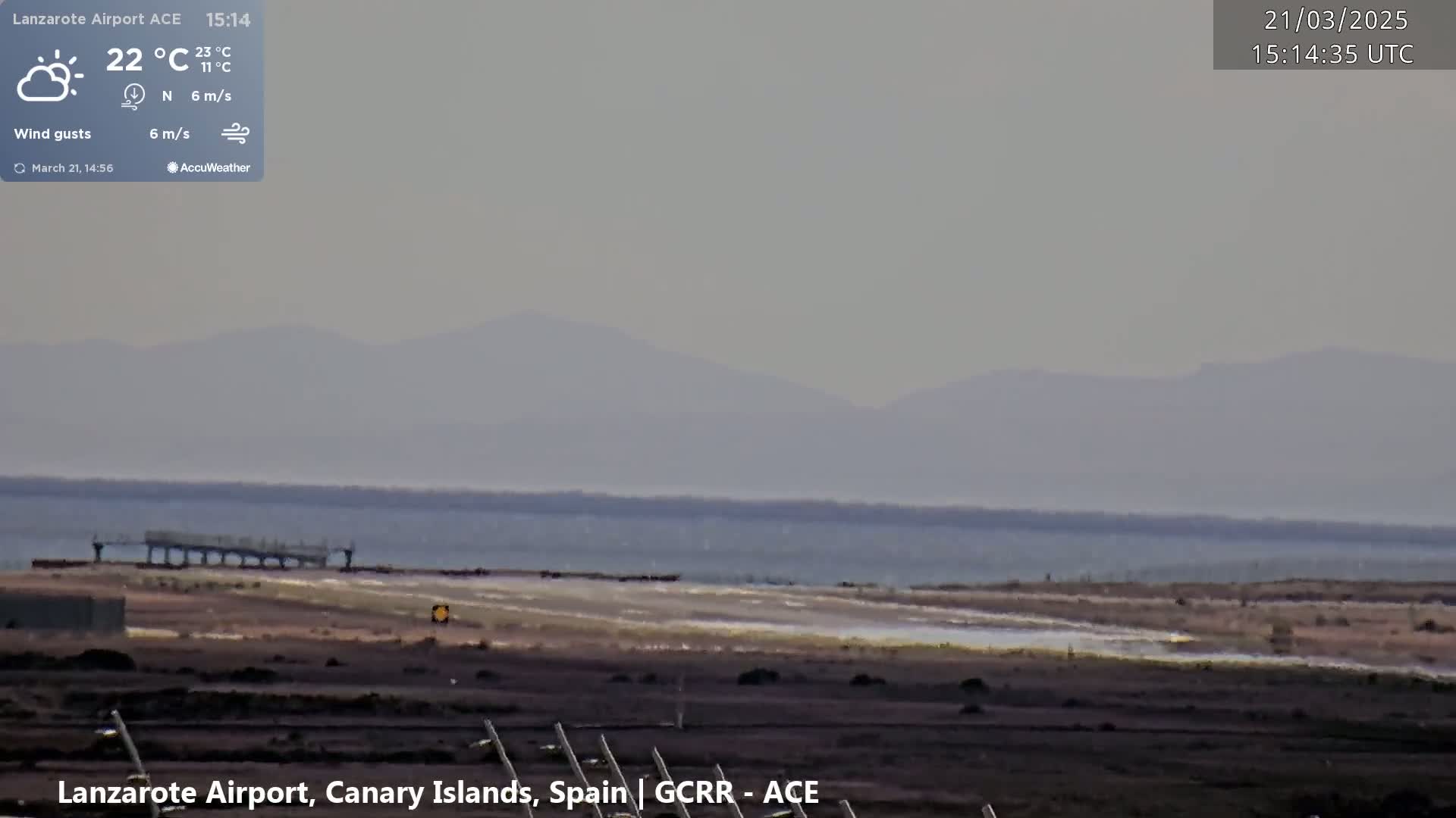 A hazy, partly sunny view of a coastal airport with a calm body of water and distant mountains.